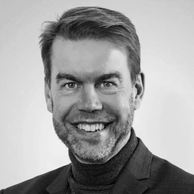 Black and white headshot of a smiling man with short hair, a beard, wearing a dark blazer and turtleneck against a plain background.