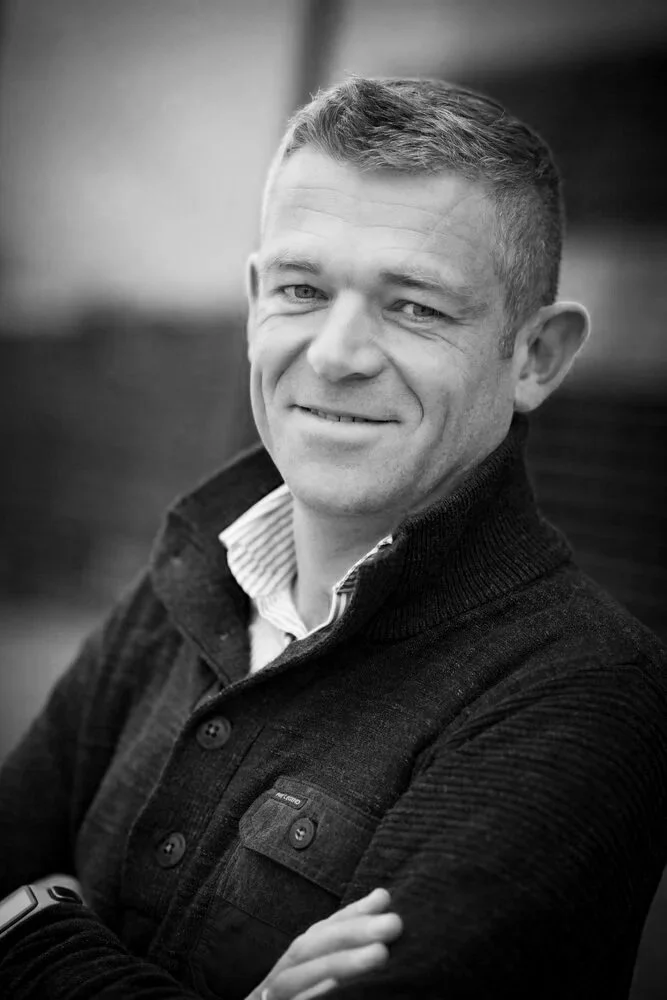 Black and white portrait of a man smiling with crossed arms. He has short hair, wearing a collared shirt under a dark sweater with a zip collar.