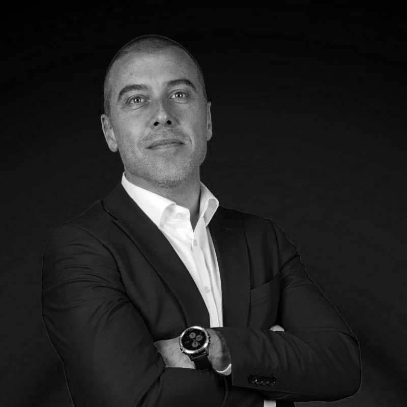 Black and white portrait of a man in a suit with arms crossed, wearing a watch, against a dark background.
