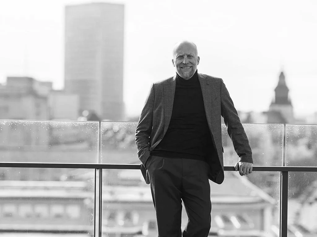 A smiling older man with a beard, wearing a suit jacket and turtleneck, standing on a balcony with a city skyline in the background.