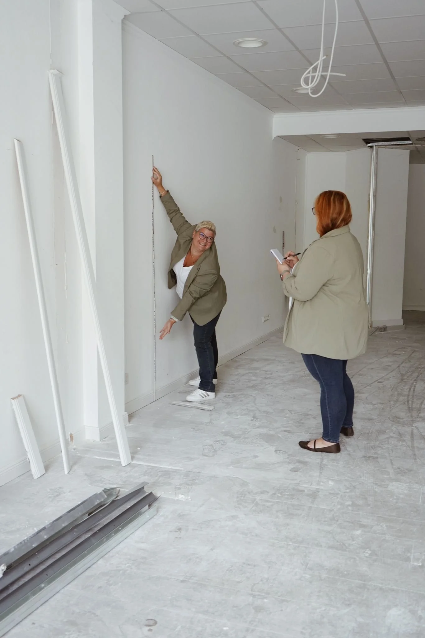 Zwei Frauen bei Messarbeiten in einem Raum im Renovierungsprozess, eine misst die Wandhöhe, die andere macht Notizen auf einem notizblock.