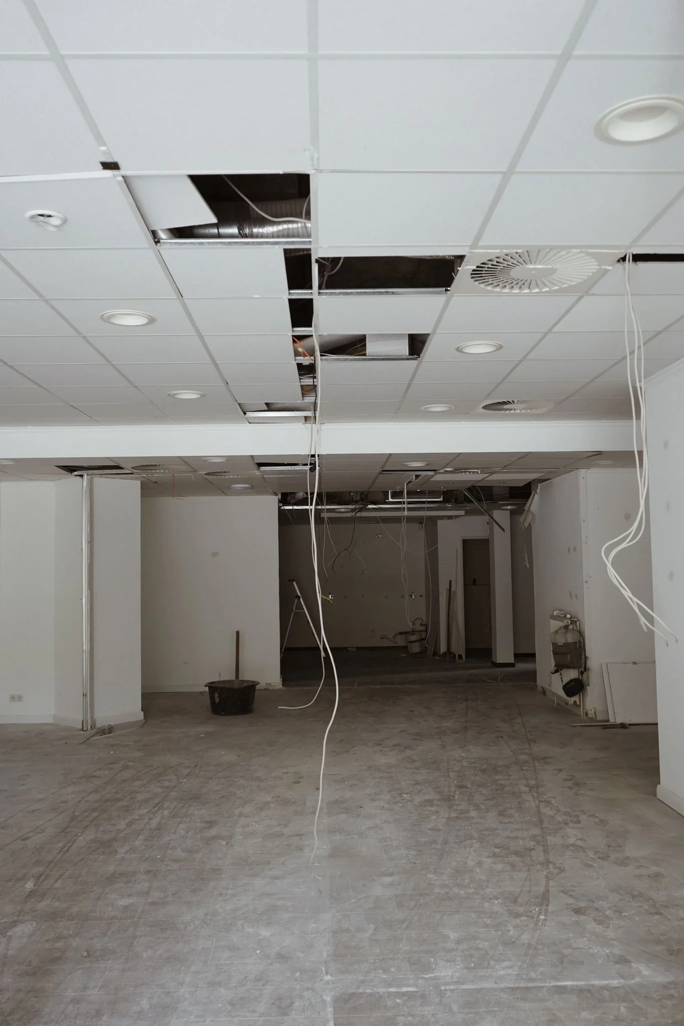 Bürorenovierungsarbeiten mit offenem Deckenbereich, sichtbaren Kabeln und unvollendeter Decke, Staub und Bauutensilien auf dem Boden.