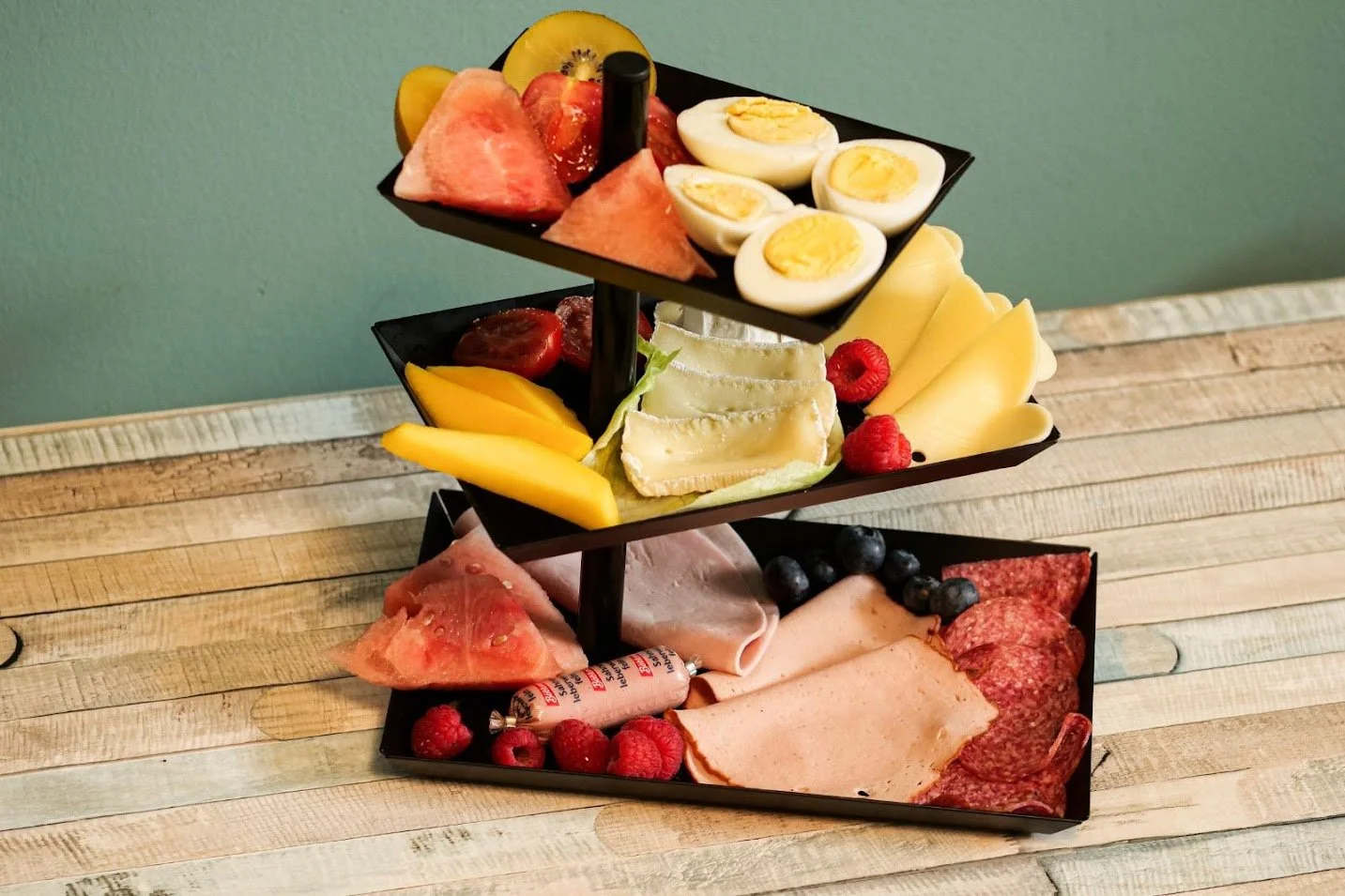 Dreistöckiges Käse- und Wurstplatte mit Früchten, gekochten Eiern, Käse, Salami, Schinken, Blaubeeren, Himbeeren, Wassermelone, Honig, und verschiedenen Käsesorten auf einem Holztisch.