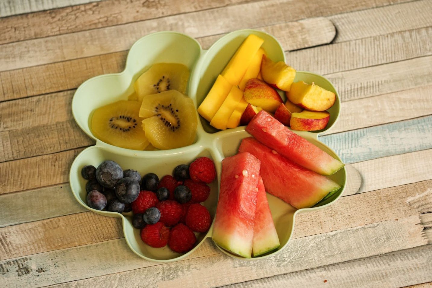 Eine Schüssel in Form einer Blume mit verschiedenen Früchten, inklusive Kiwi, Mango, Erdbeeren, Blaubeeren, Wassermelone, Pfirsich und Apfel, auf einem Holztisch.