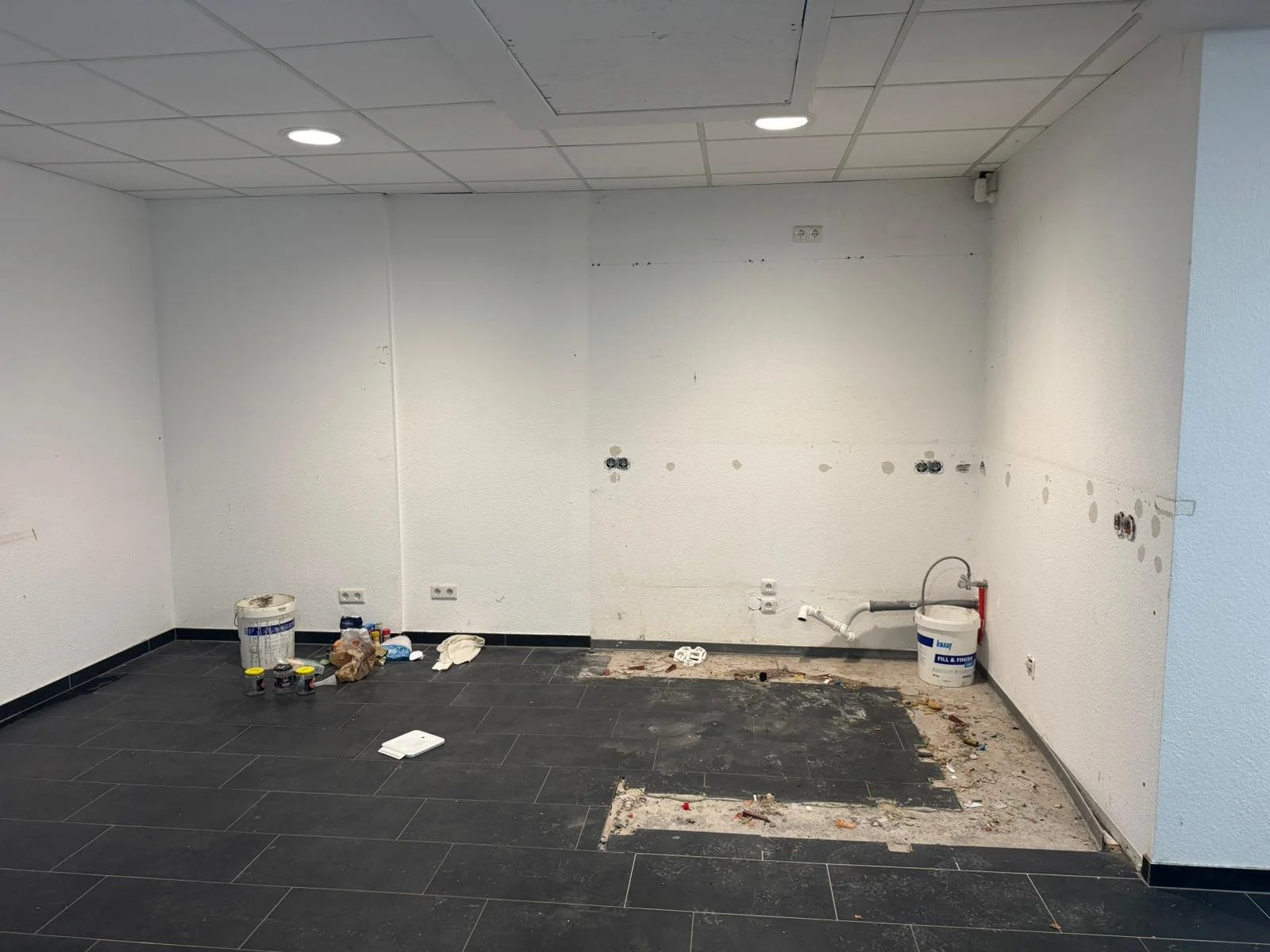 Leeres Zimmer mit dunklen Fliesen und weißen Wänden, Umbauarbeiten im Gange, sichtbare Wasseranschlüsse und Baumaterialien auf dem Boden.