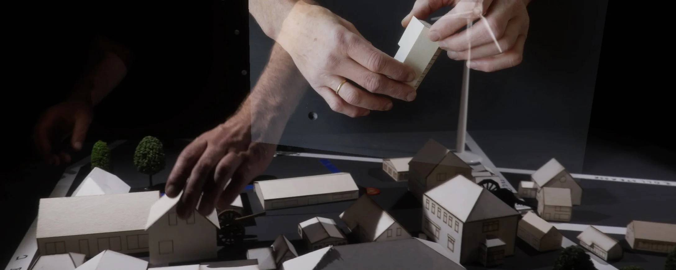 Hands of interviewees holding miniature houses in a video shown in AR above a model where a performer works with these same miniature houses.