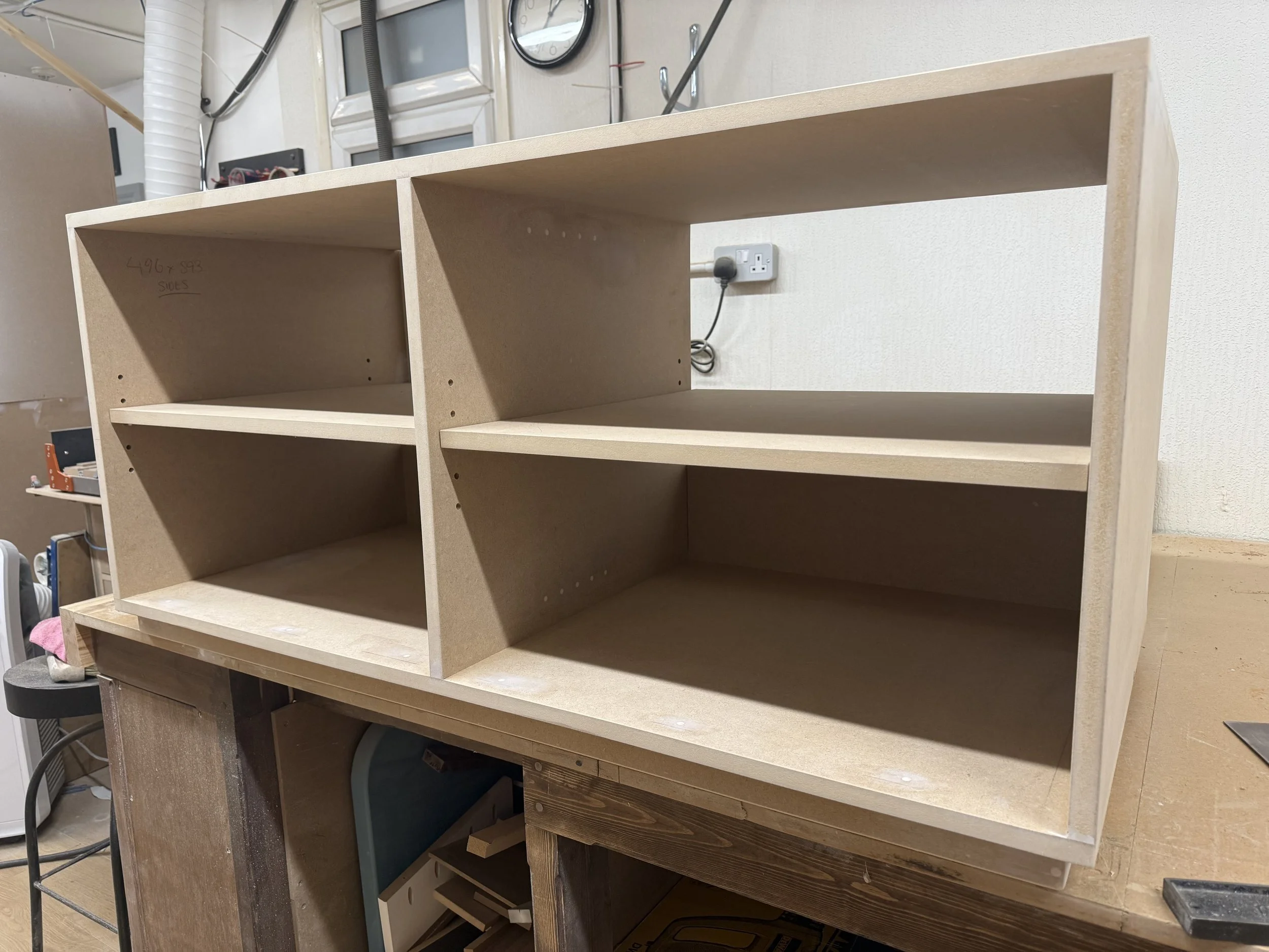 unfinished wooden shelving unit with three compartments, placed on a workbench in a workshop.