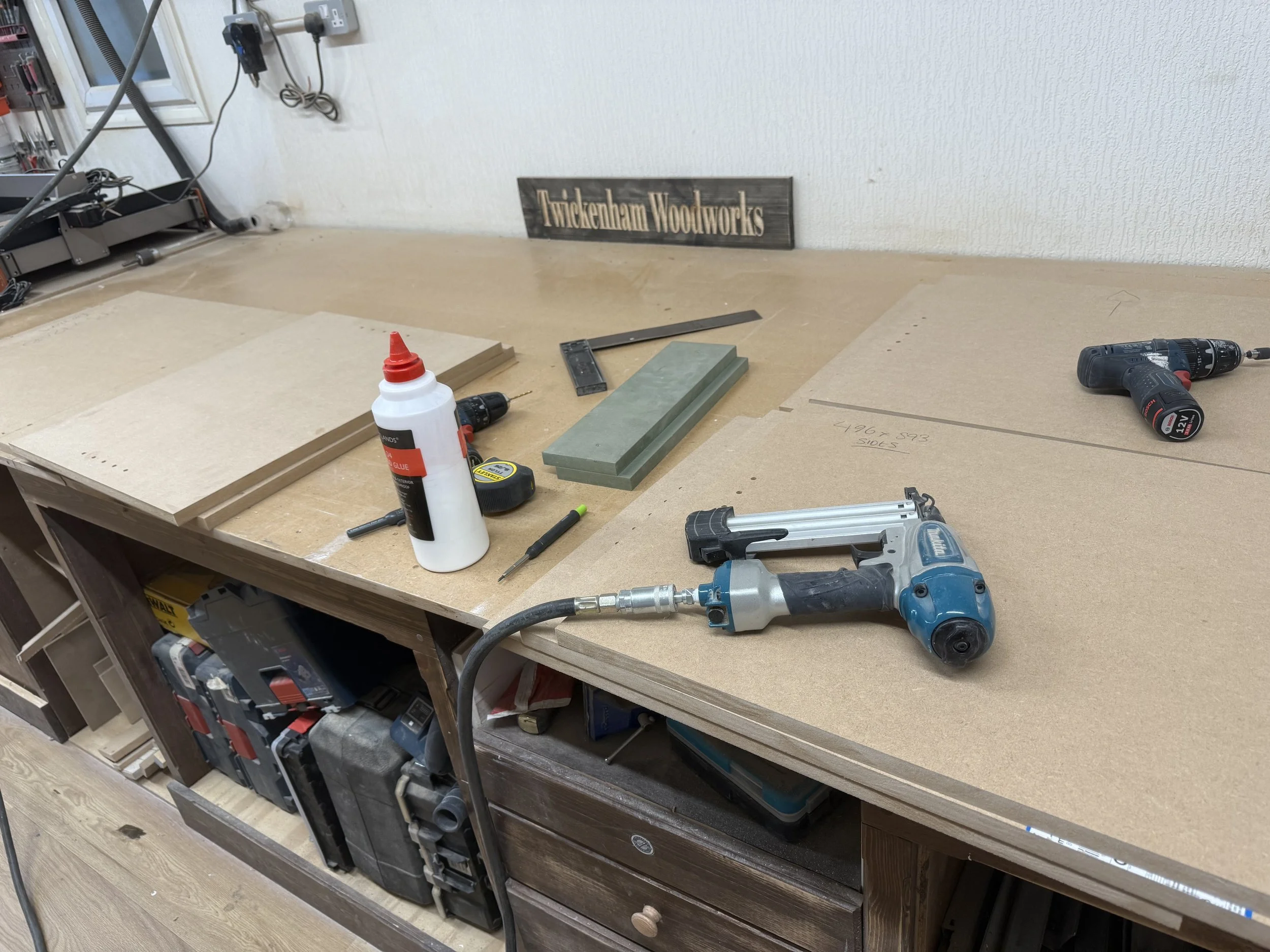 Woodworking workshop with a workbench holding a cordless drill, a nail gun, a bottle of glue, and various wooden pieces; tools and supplies are organized nearby, and a sign reading 'Twickenham Woodworks' is visible on the wall.