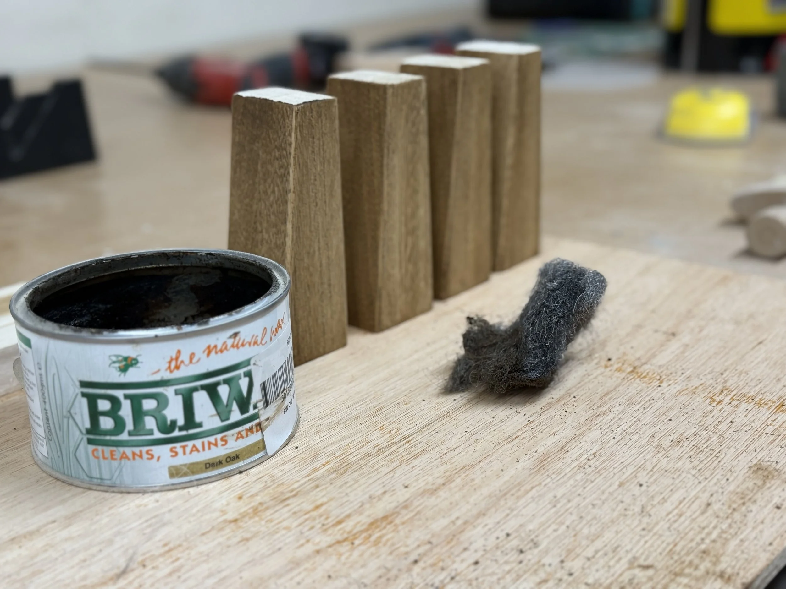 A woodworking workspace with a can of Briwax wood finish, five vertical wooden blocks, and a gray scouring pad on a wooden surface.