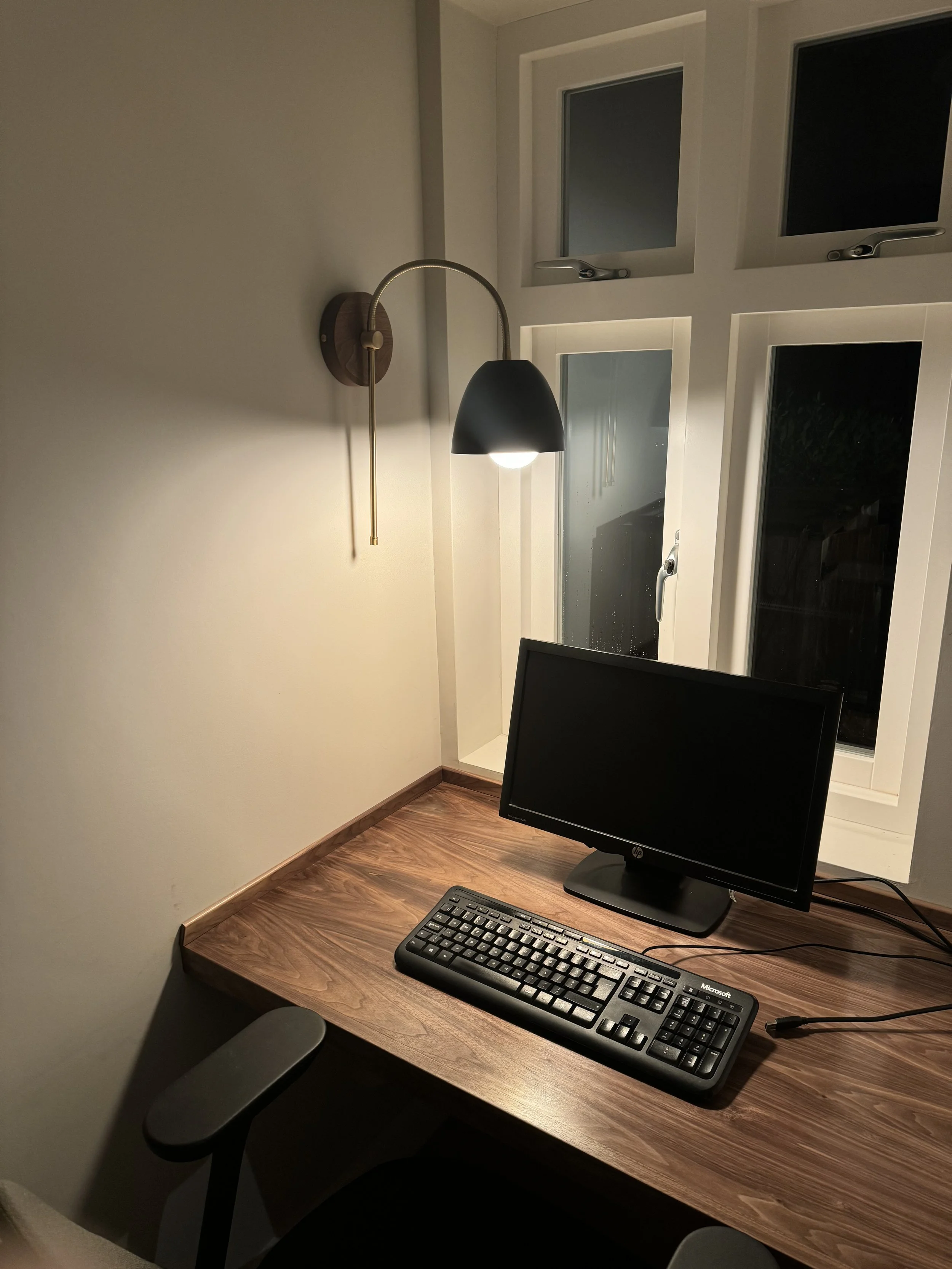 A home office setup with a wooden desk, a computer monitor, keyboard, and a desk chair. There's a wall-mounted lamp near the window, which is closed, at night.