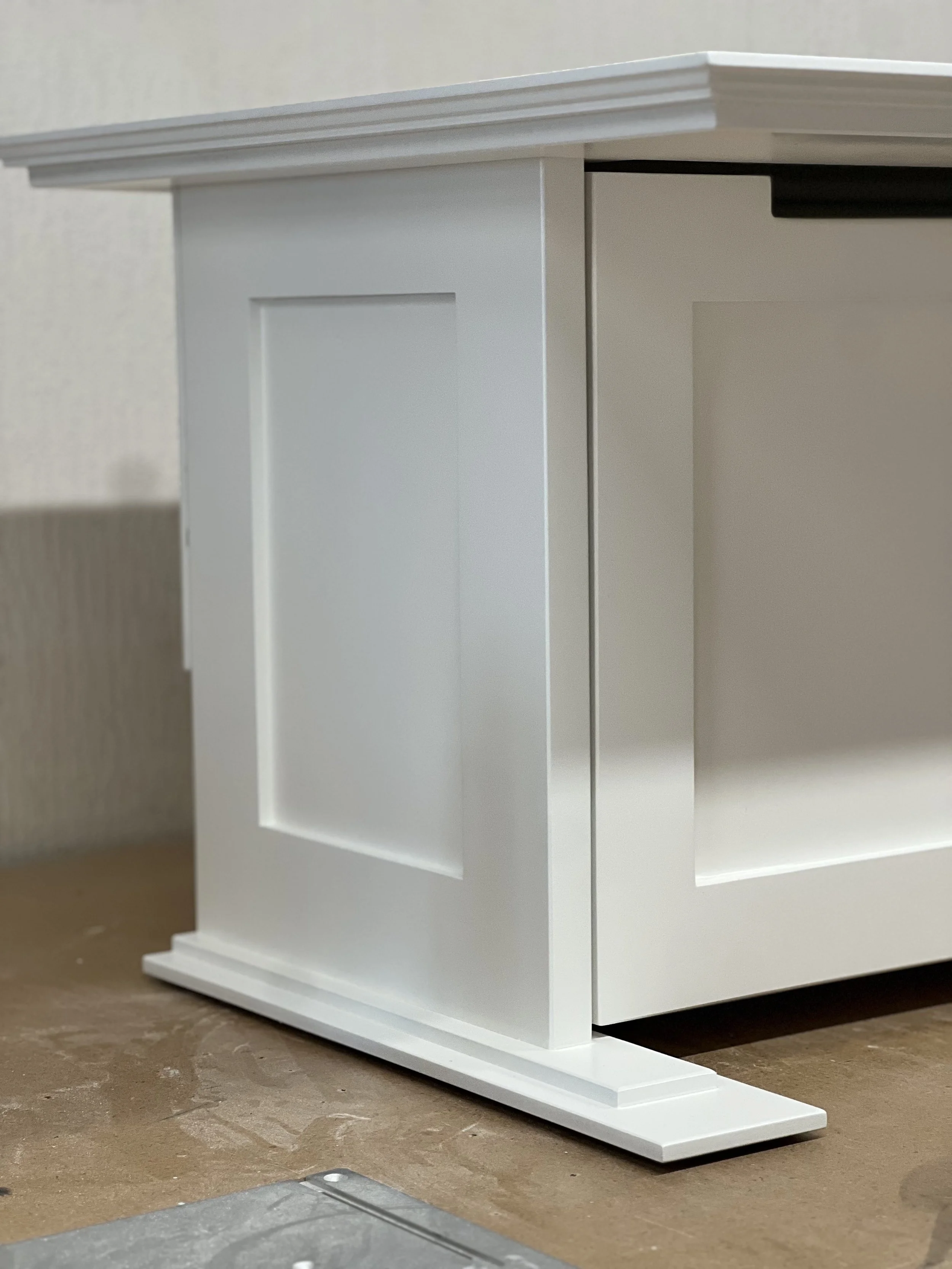 Close-up of a white wooden cabinet with a recessed panel door and a decorative base, placed on a wooden surface.