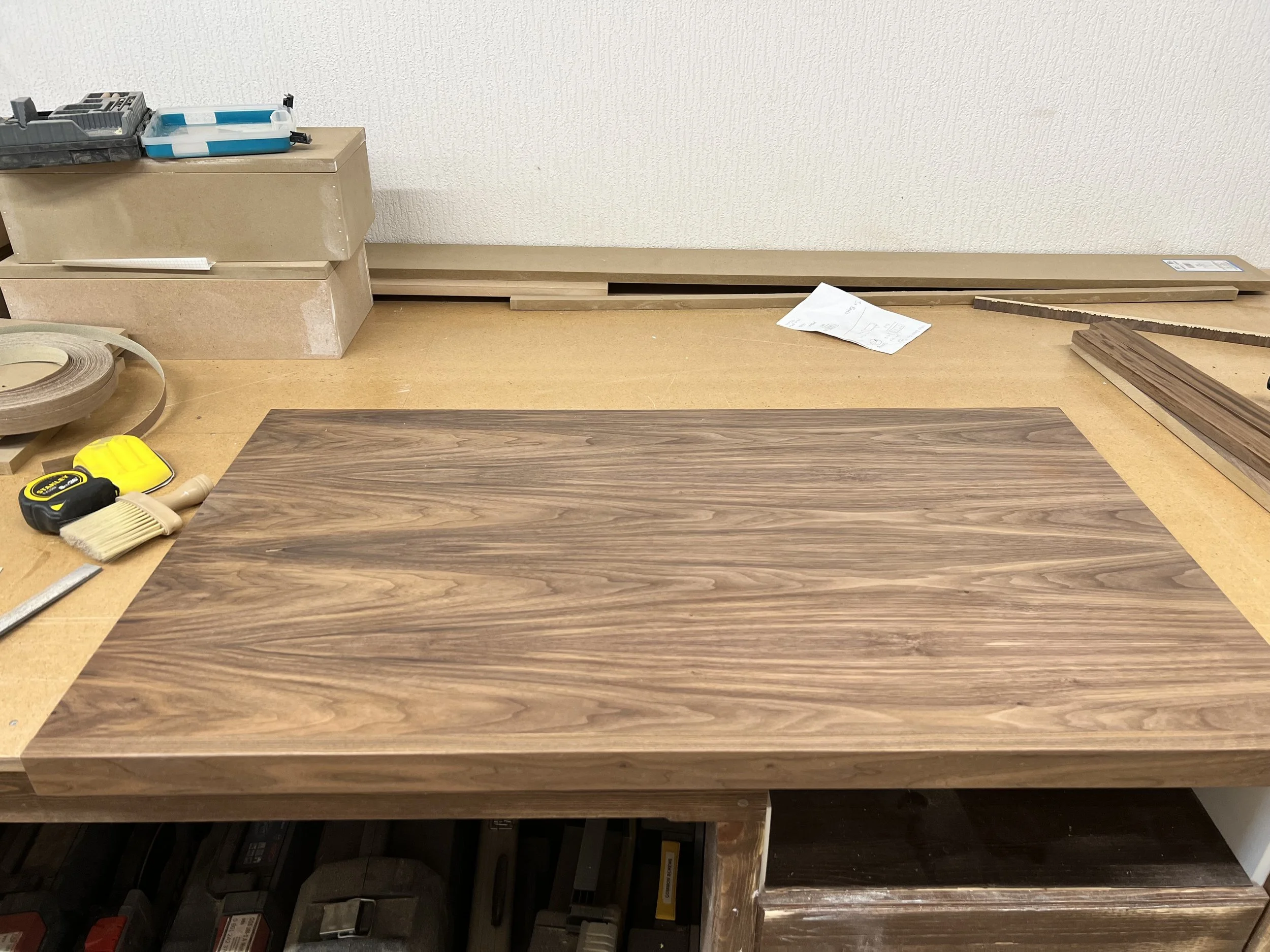 A workshop workbench with a large wood panel, tools, and hardware, with wood pieces and supplies in the background.