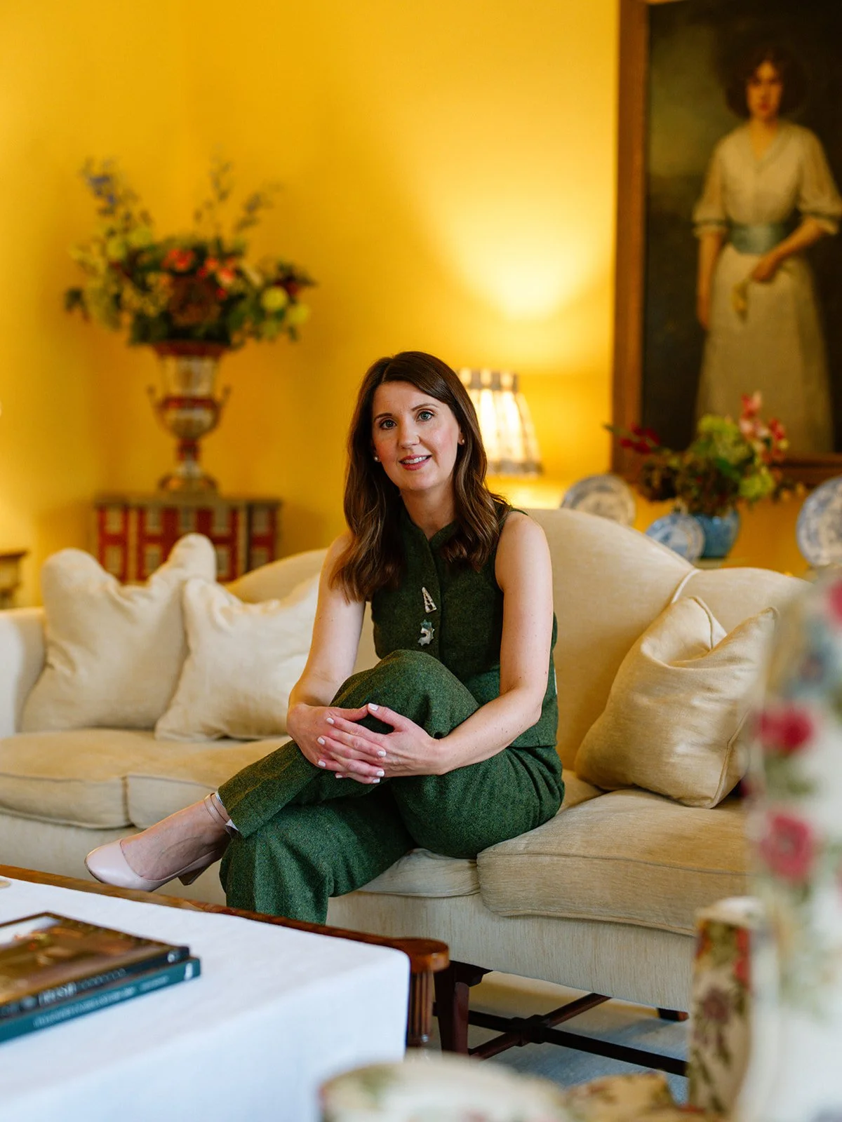 A woman with shoulder-length brown hair wearing a sleeveless dark green top and matching green pants sits on a cream-colored sofa with her hands clasped around her knee. The room has yellow walls, a large floral arrangement and a painting of a woman in a cream dress hanging behind her, and a small table with a book and a floral-patterned chair in the foreground.