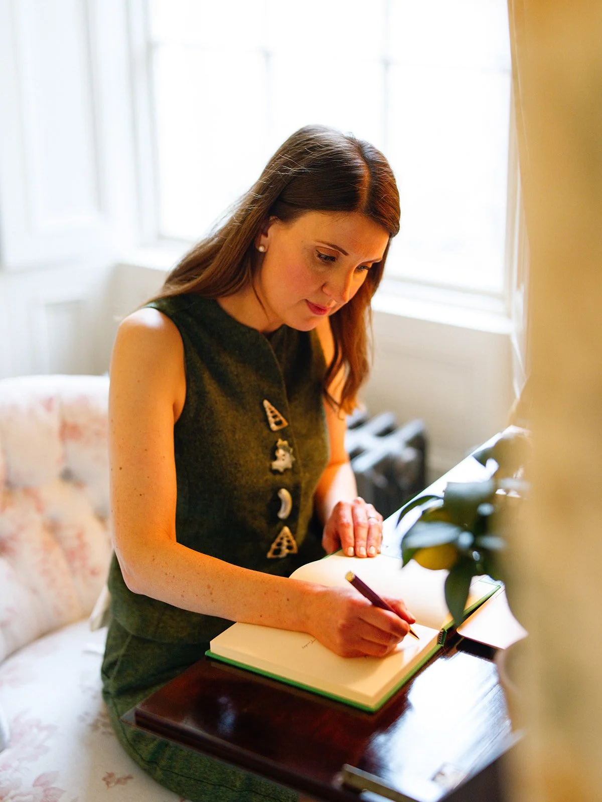 A woman with long brown hair sitting at a wooden table, writing in a notebook near a window with white units, wearing a dark sleeveless top with decorative pins.