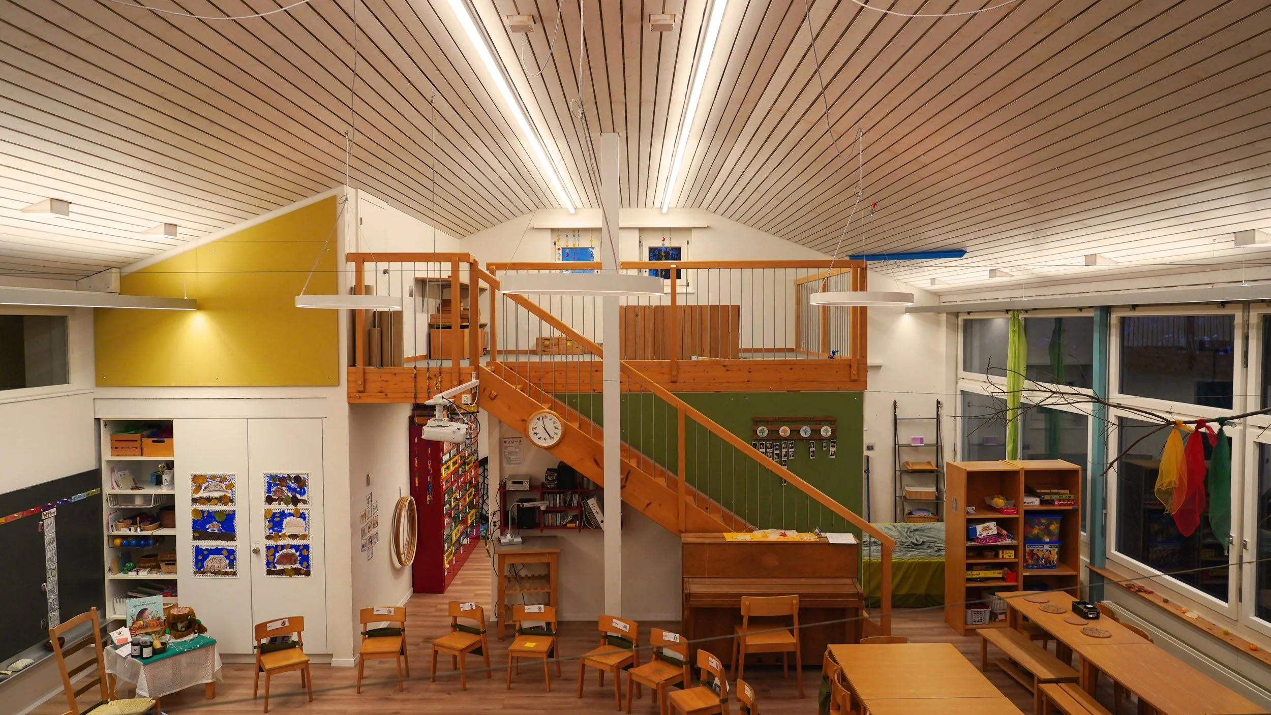 Interior view of a preschool or classroom with wooden furniture, a staircase, and colorful decorations. Multilayered illumination with different light sources.