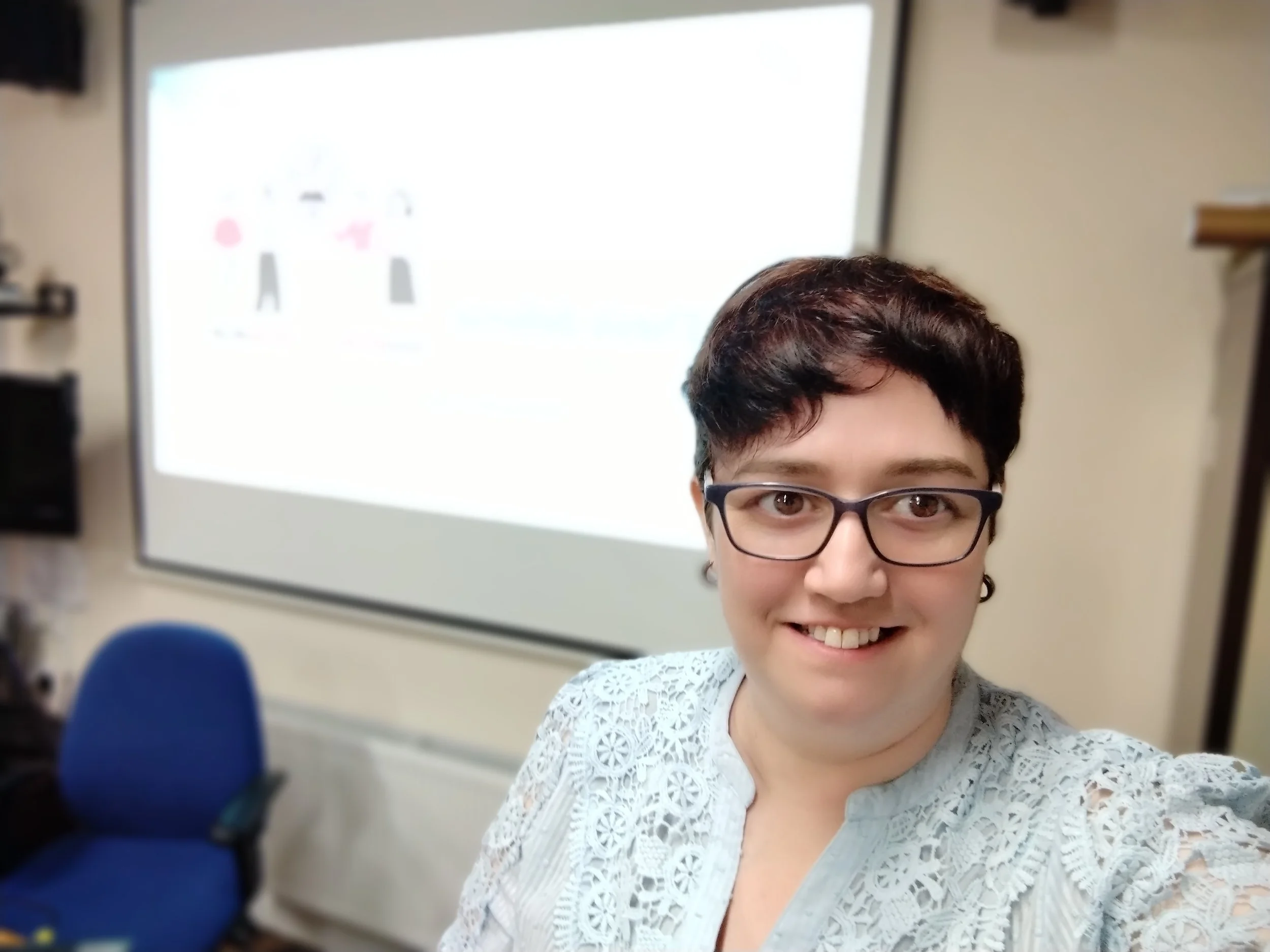 A woman taking a selfie in a classroom or office setting with a large whiteboard or screen behind her, wearing glasses and a lace top, smiling at the camera.