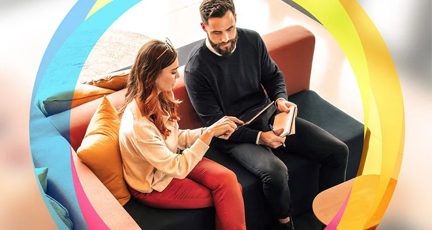 Man and woman sitting on a sofa looking at a tablet together in a brightly lit room with colorful abstract graphic overlay.