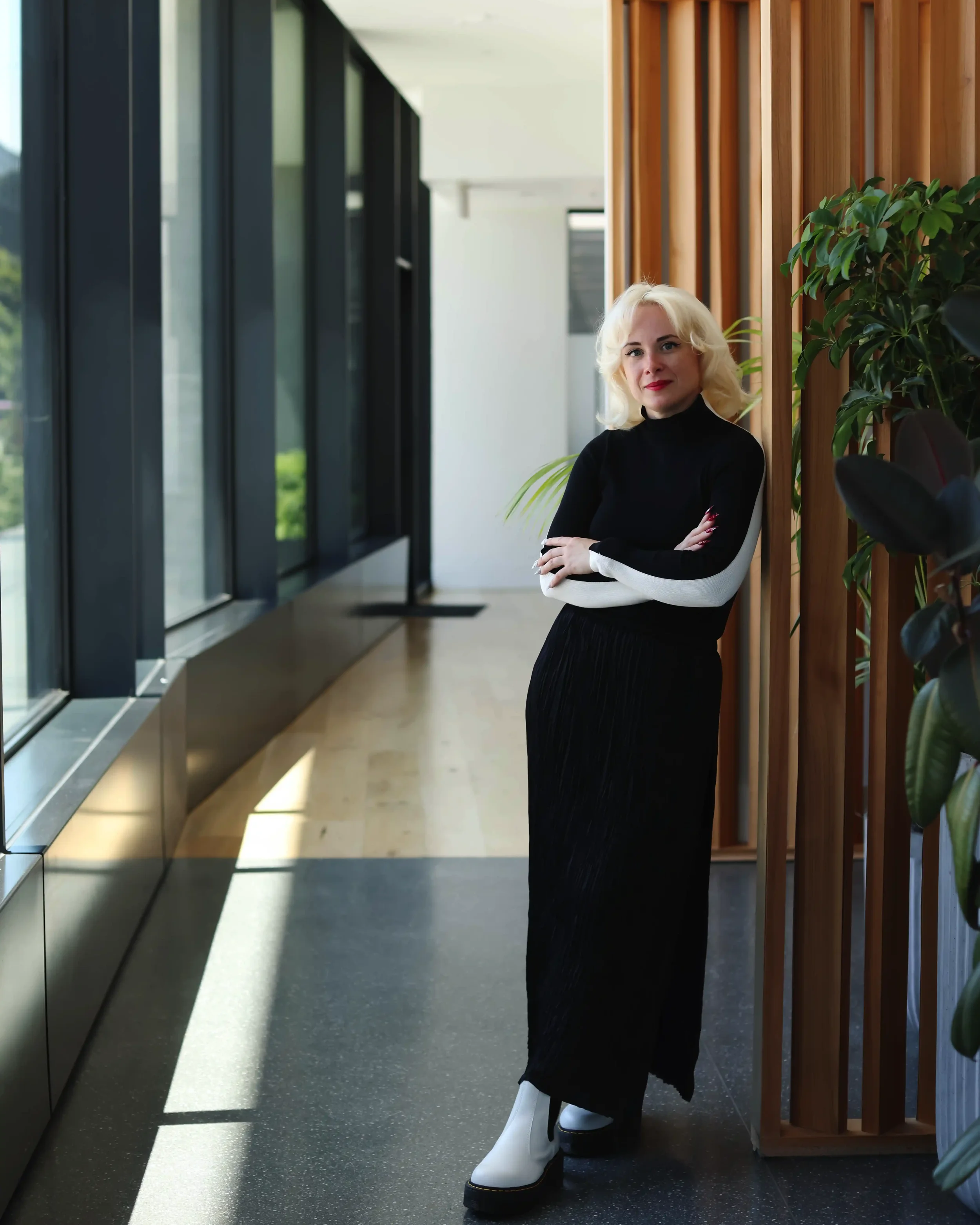 A woman standing in a modern corridor with large windows, wooden wall panels, and potted plants.