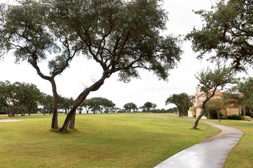 A park with green grass, several trees, and a winding concrete path, with a house visible in the background under a cloudy sky.
