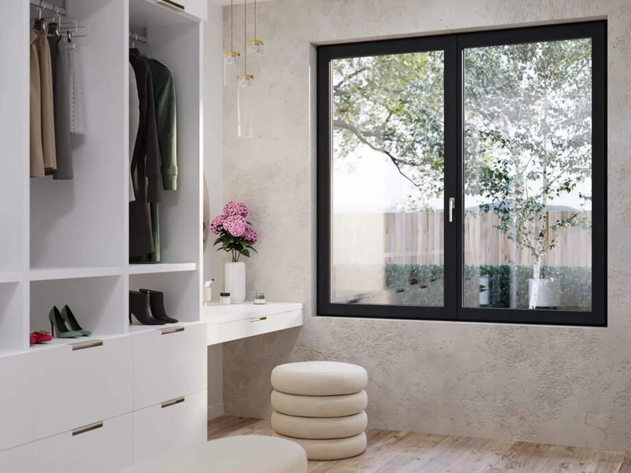 A modern bedroom with a large window showing trees outside, white closet with hanging clothes, a white desk with pink flowers, and a white rounded stool.