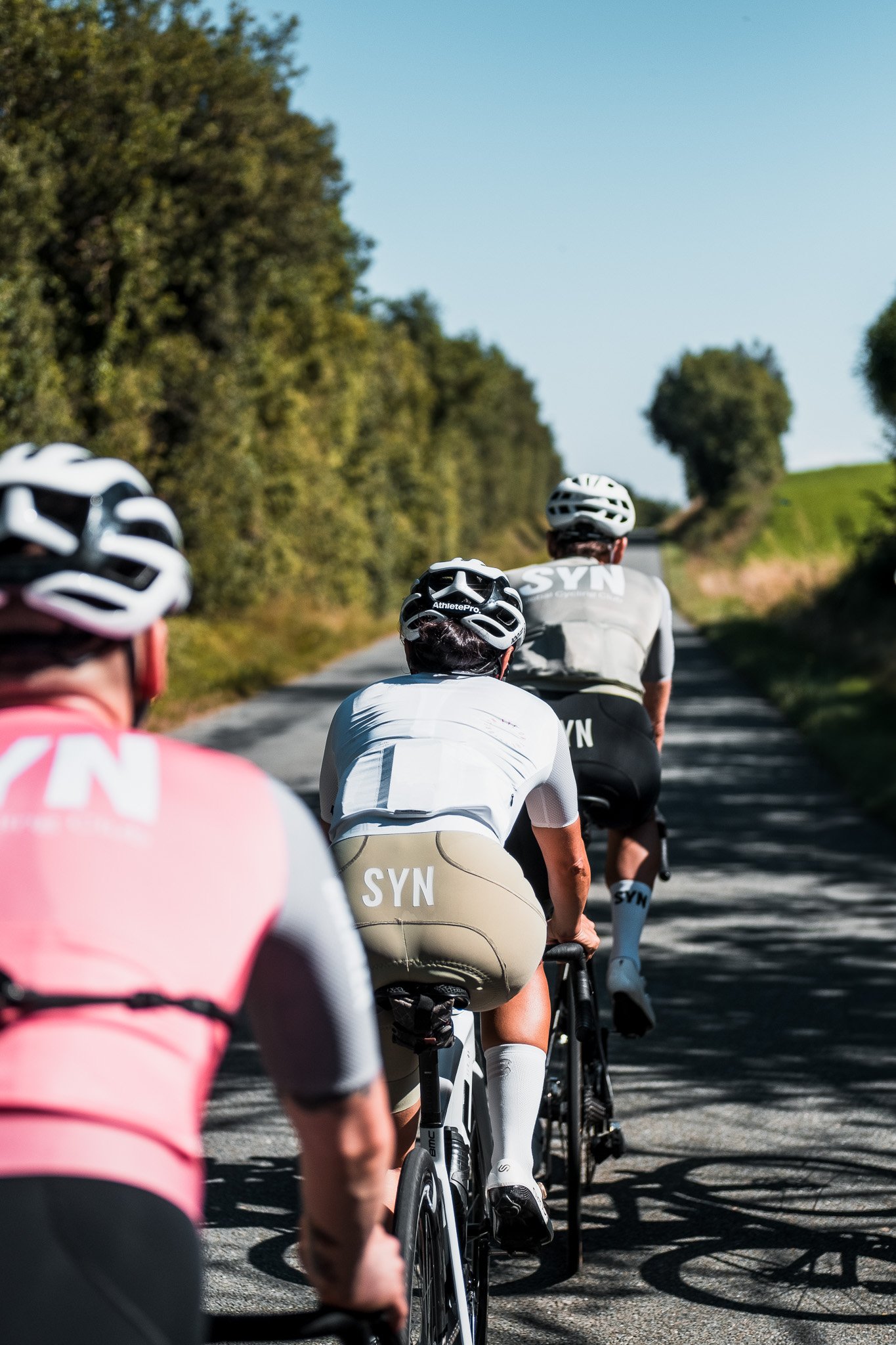 A group of cyclists riding their bikes on a country road on a sunny day, surrounded by green trees and grass.