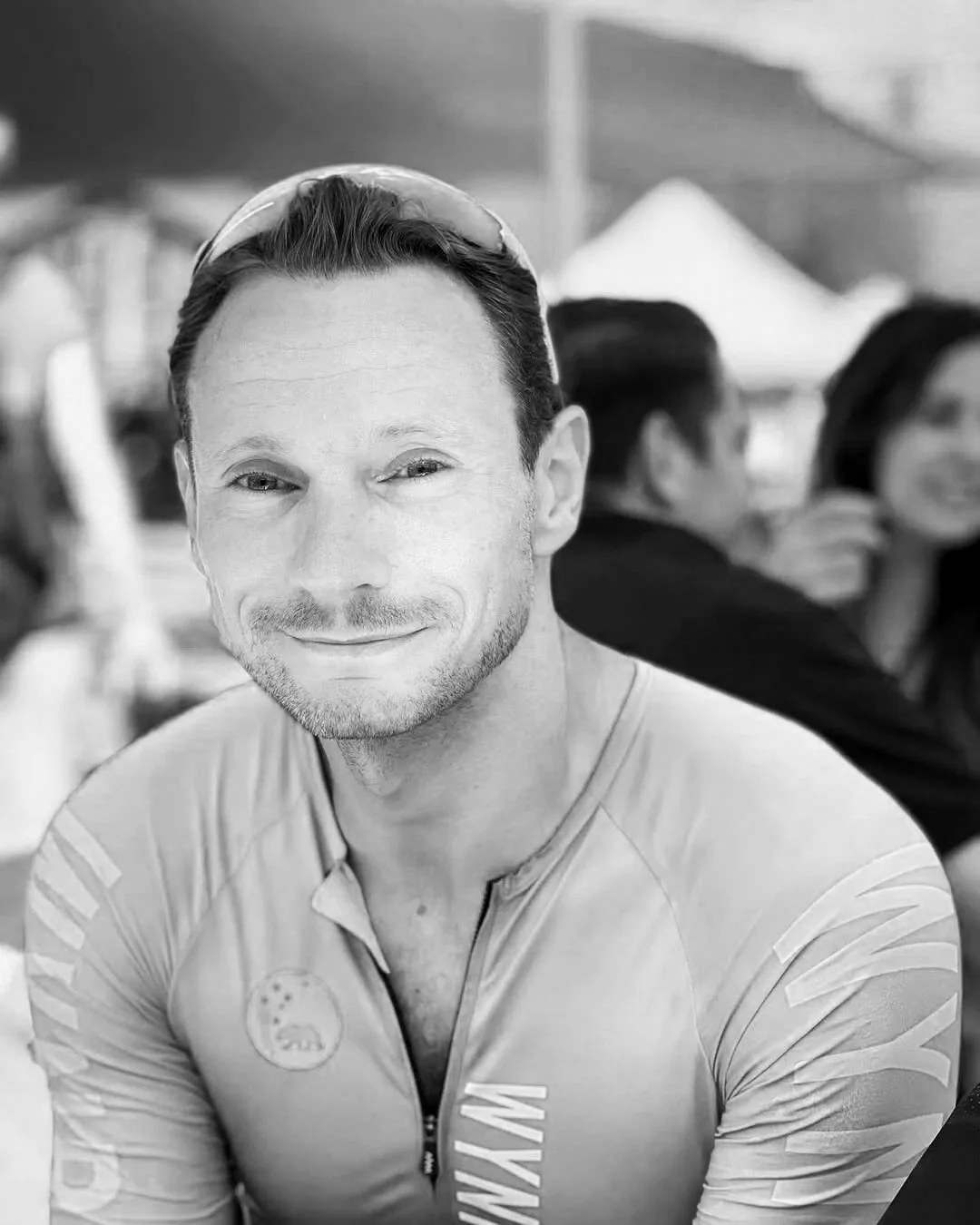 A smiling man wearing a cycling jersey, sunglasses on his head, with a blurred background showing other people and tents at an outdoor event.