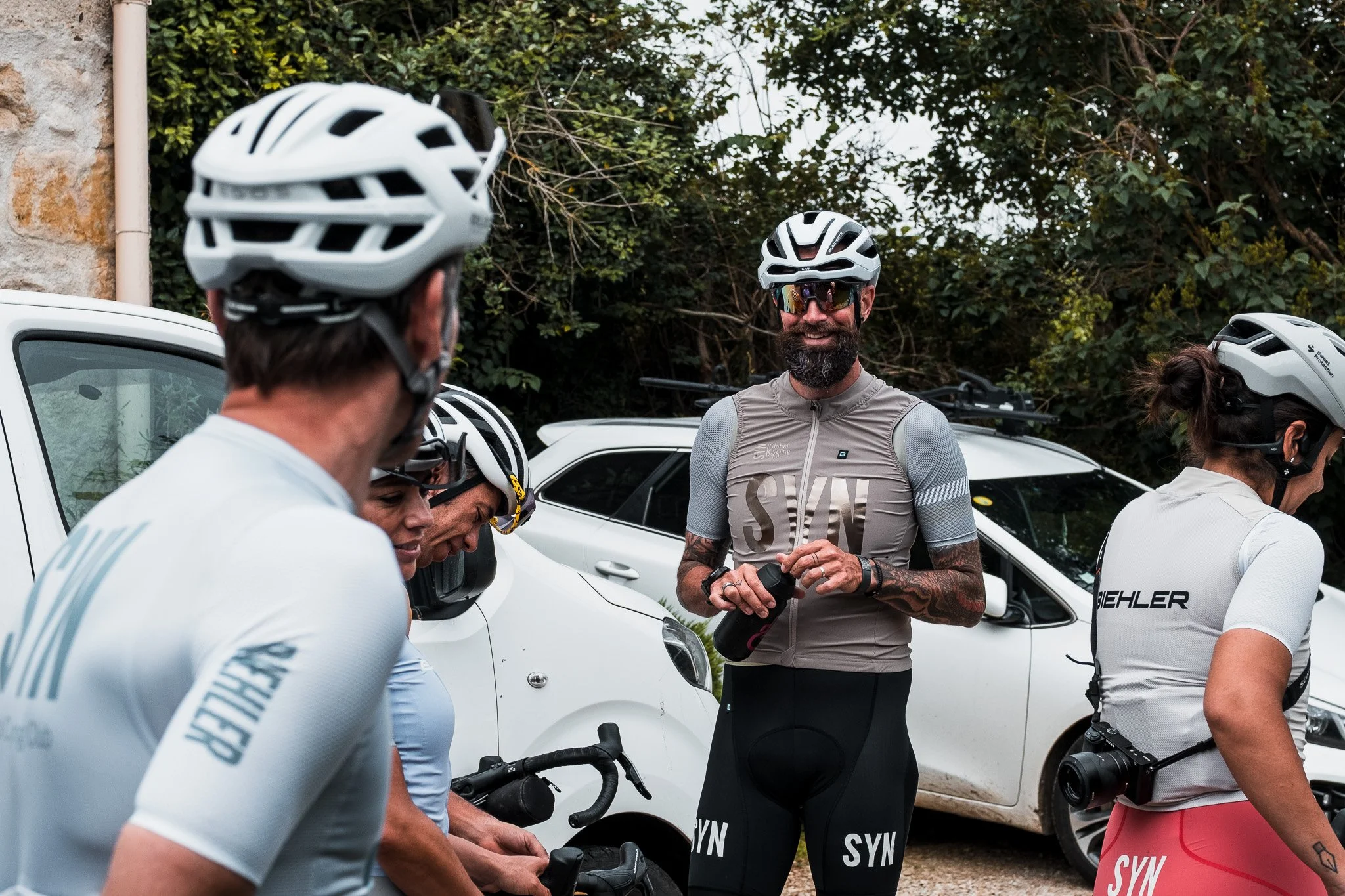A group of cyclists wearing helmets and cycling gear, gathered outdoors near parked cars and trees, engaged in conversation.
