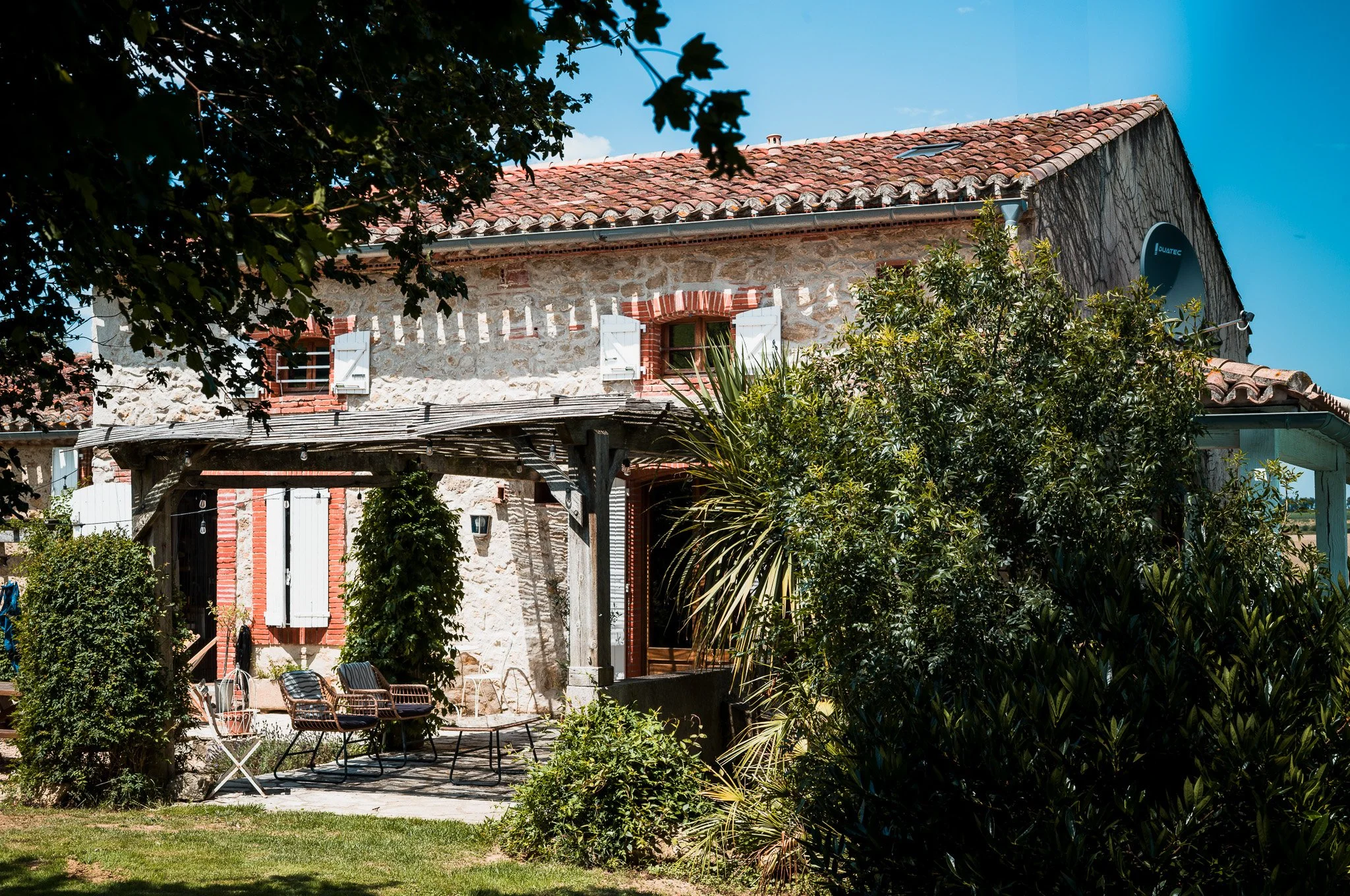 Stone house with red tile roof and open shutters, surrounded by greenery and outdoor furniture.