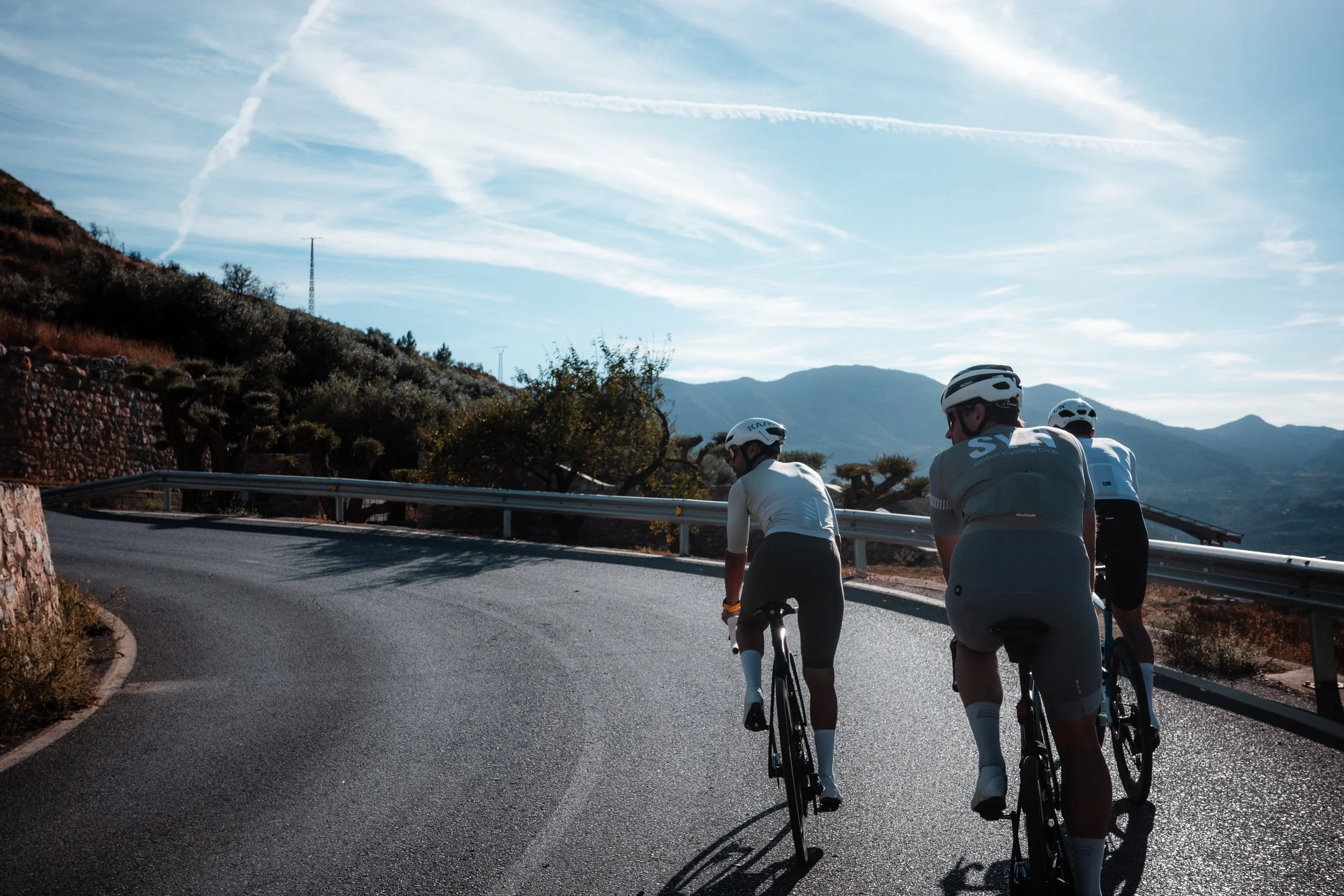 Three cyclists riding on a mountain road with scenic mountain views and a partly cloudy sky.