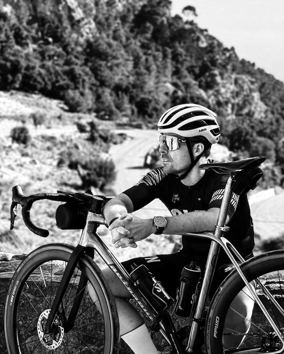 A man wearing a helmet and sunglasses sits on a racing bicycle on a mountainous trail.