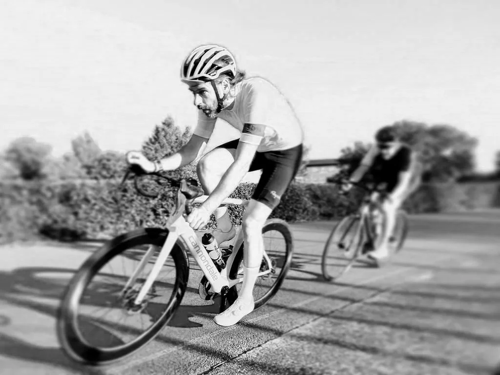 Two men riding bicycles on a paved path with trees and bushes in the background, one wearing a helmet and the other with sunglasses, both leaning forward as they pedal.