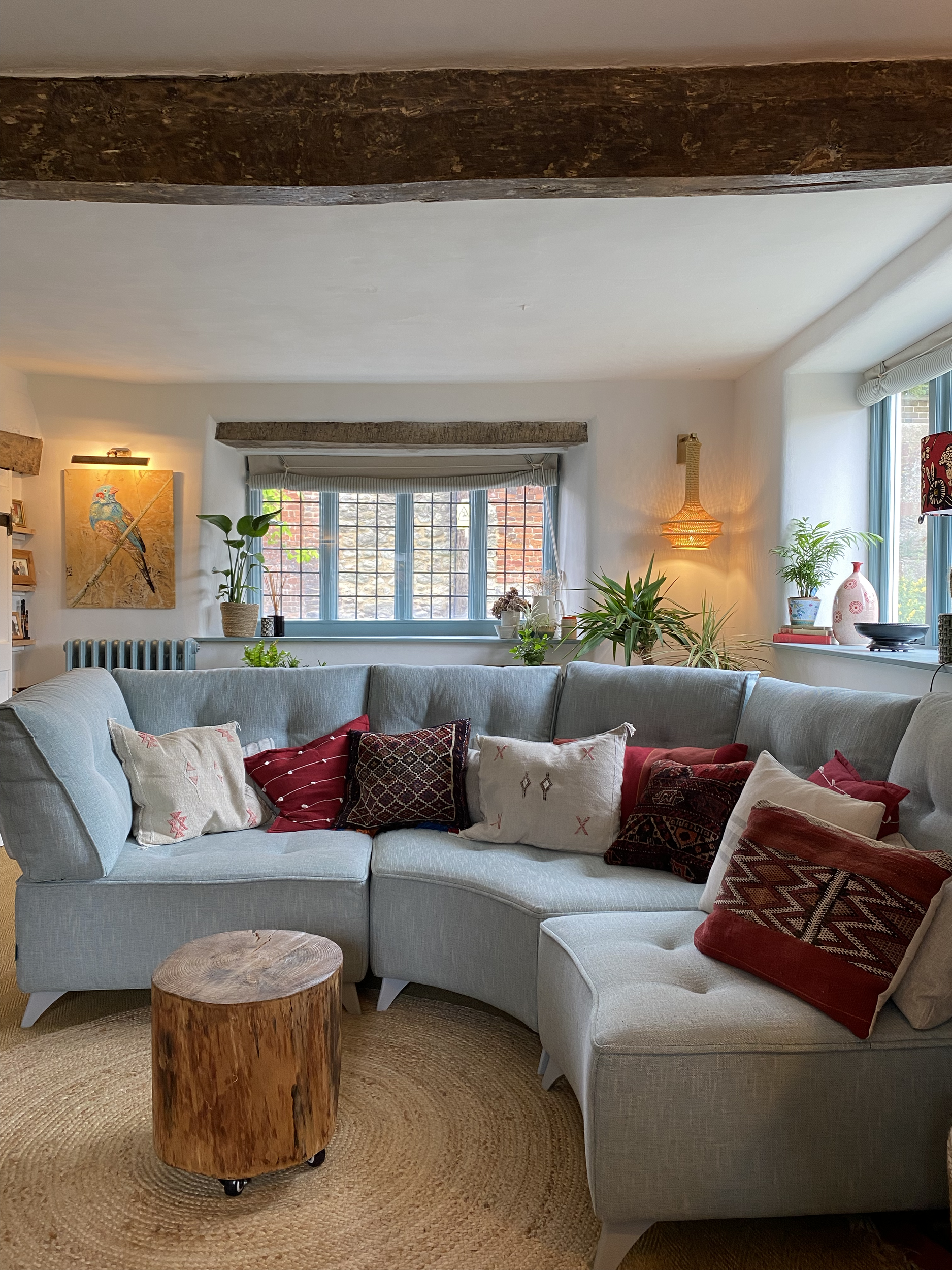 Living room with a light blue couch with various patterned pillows, a round wooden coffee table, a window with potted plants, and wall art with a bird, illuminated by warm lighting.