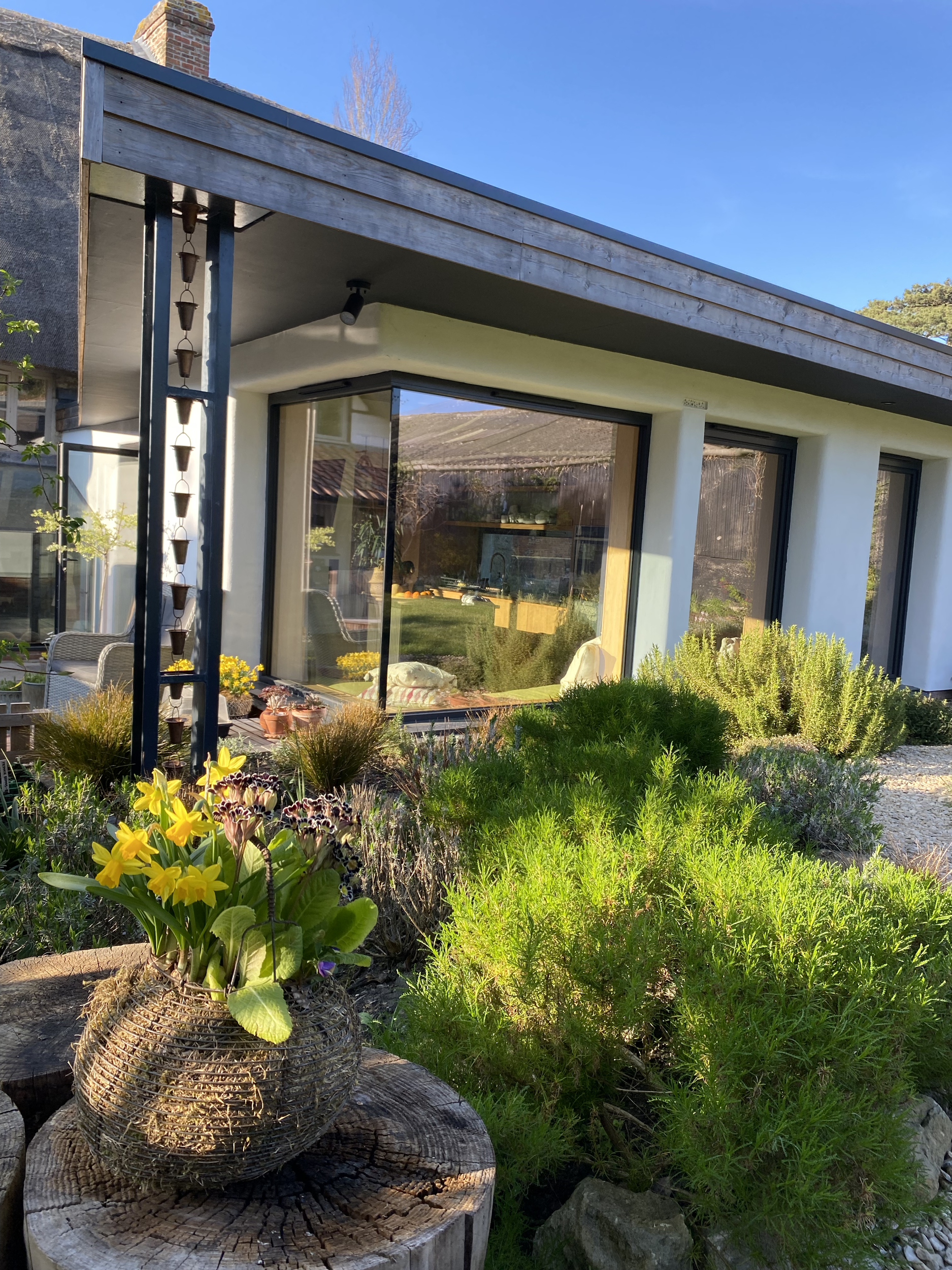 A modern house with large glass windows, a white exterior, and a patio area with potted plants, trees, and flower arrangements in a garden setting.