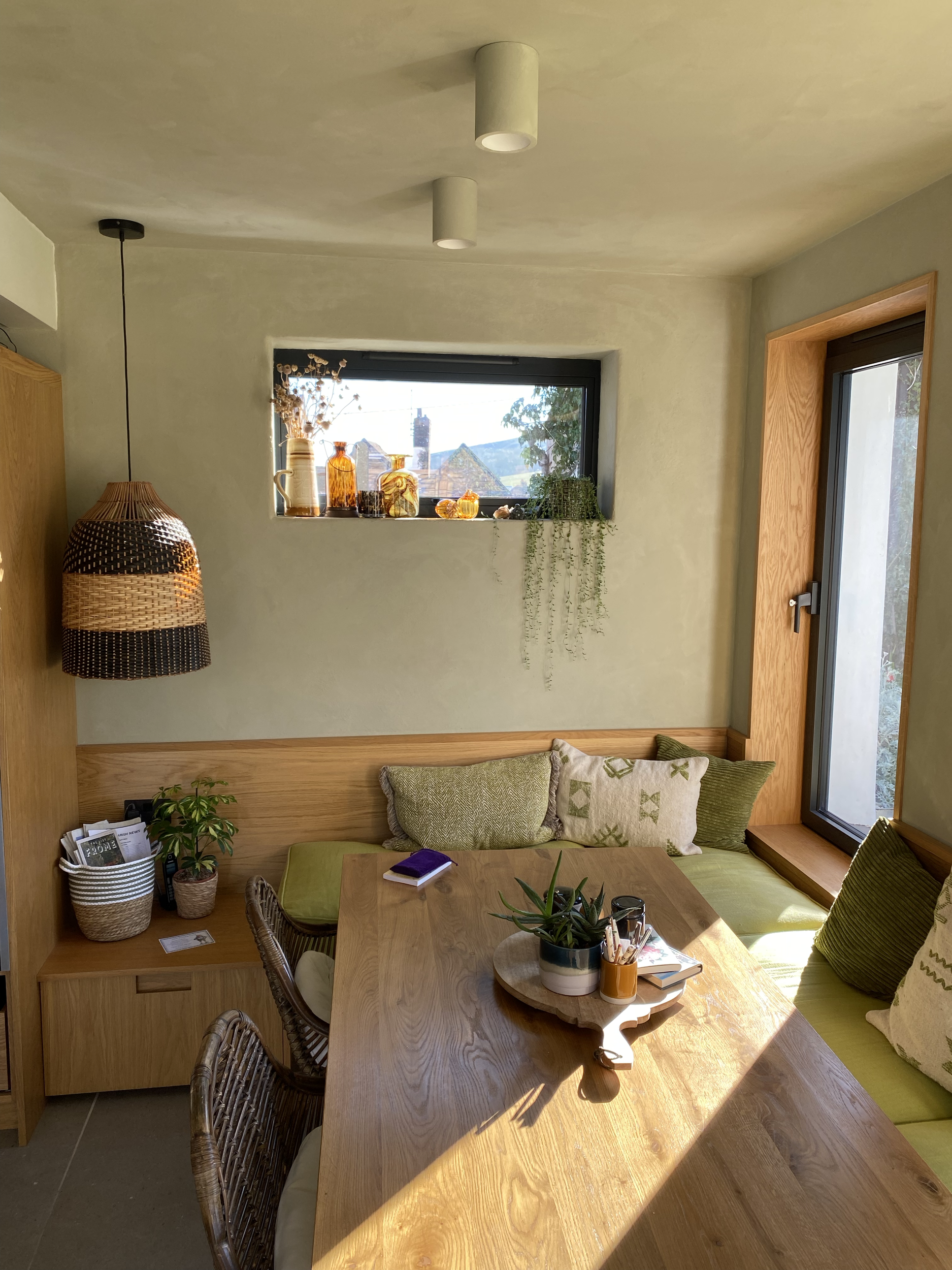 Interior view of a cozy nook with a wooden table, green cushioned seating, and decorative cushions along a wall with a window and sliding glass door, with various vases and plants on the window sill and a woven hanging light fixture.