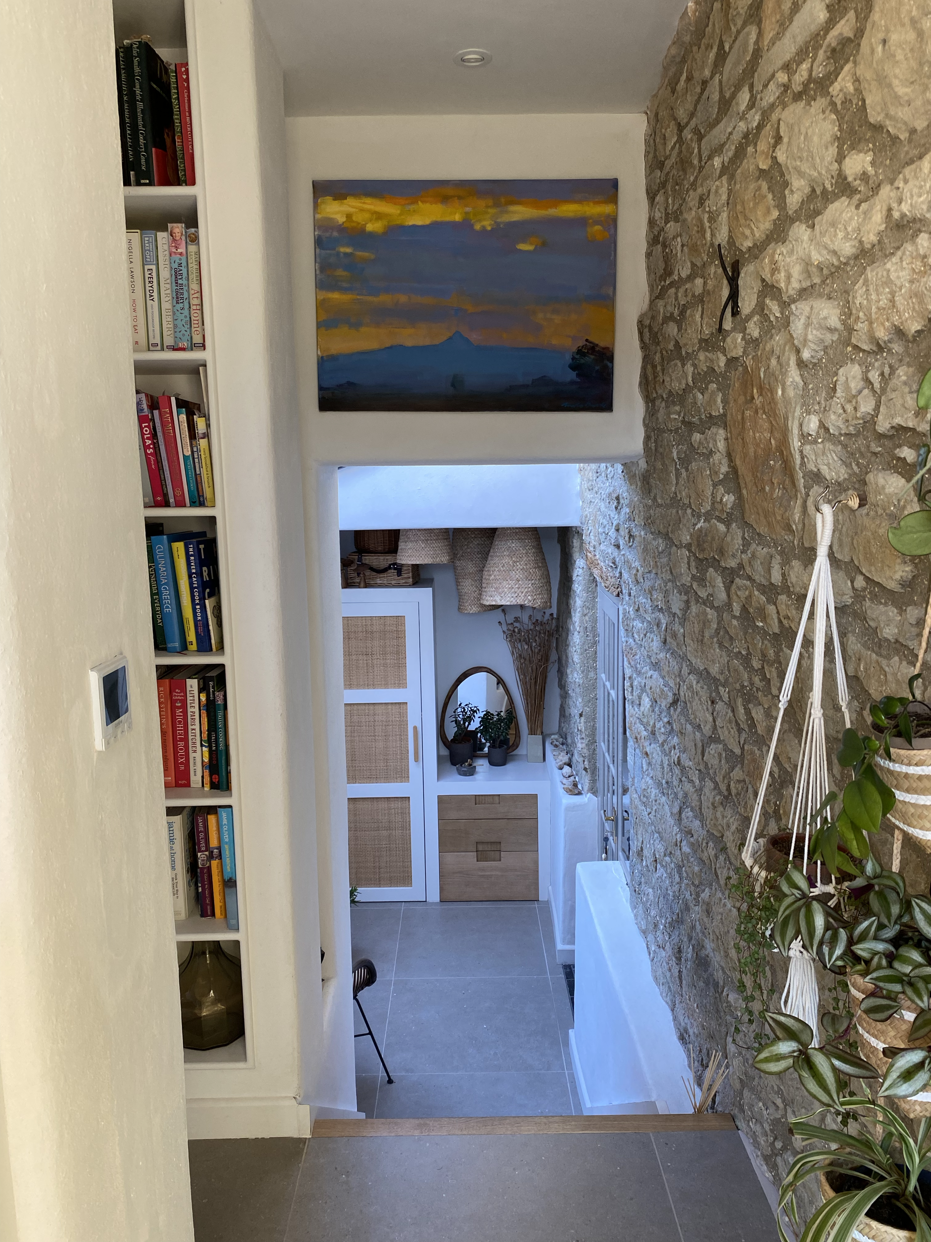 Interior view of a cozy living space with a stone wall, white cabinetry with woven rattan panels, a small wooden dresser, a mirror, potted plants, a comfortable chair, a colorful landscape painting, and a bookshelf with various books.
