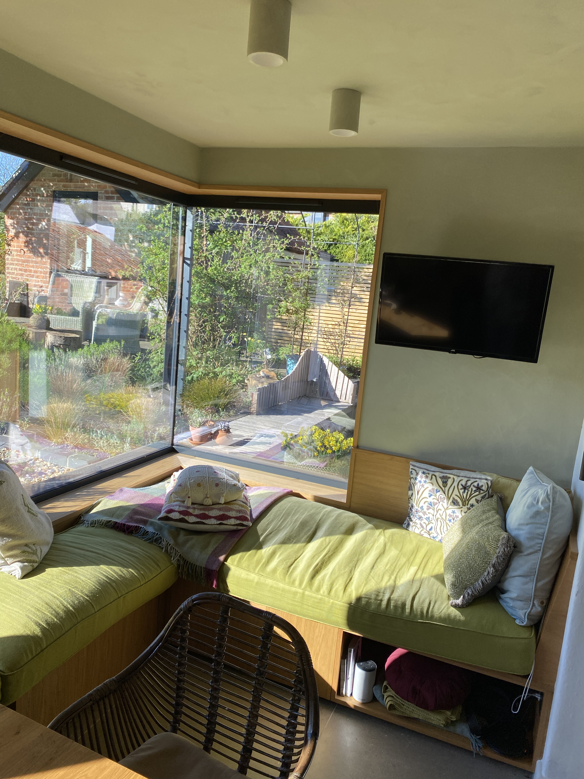 Living room with green couch, pillows, a wall-mounted TV, and a large window showing a garden with plants and outdoor furniture.