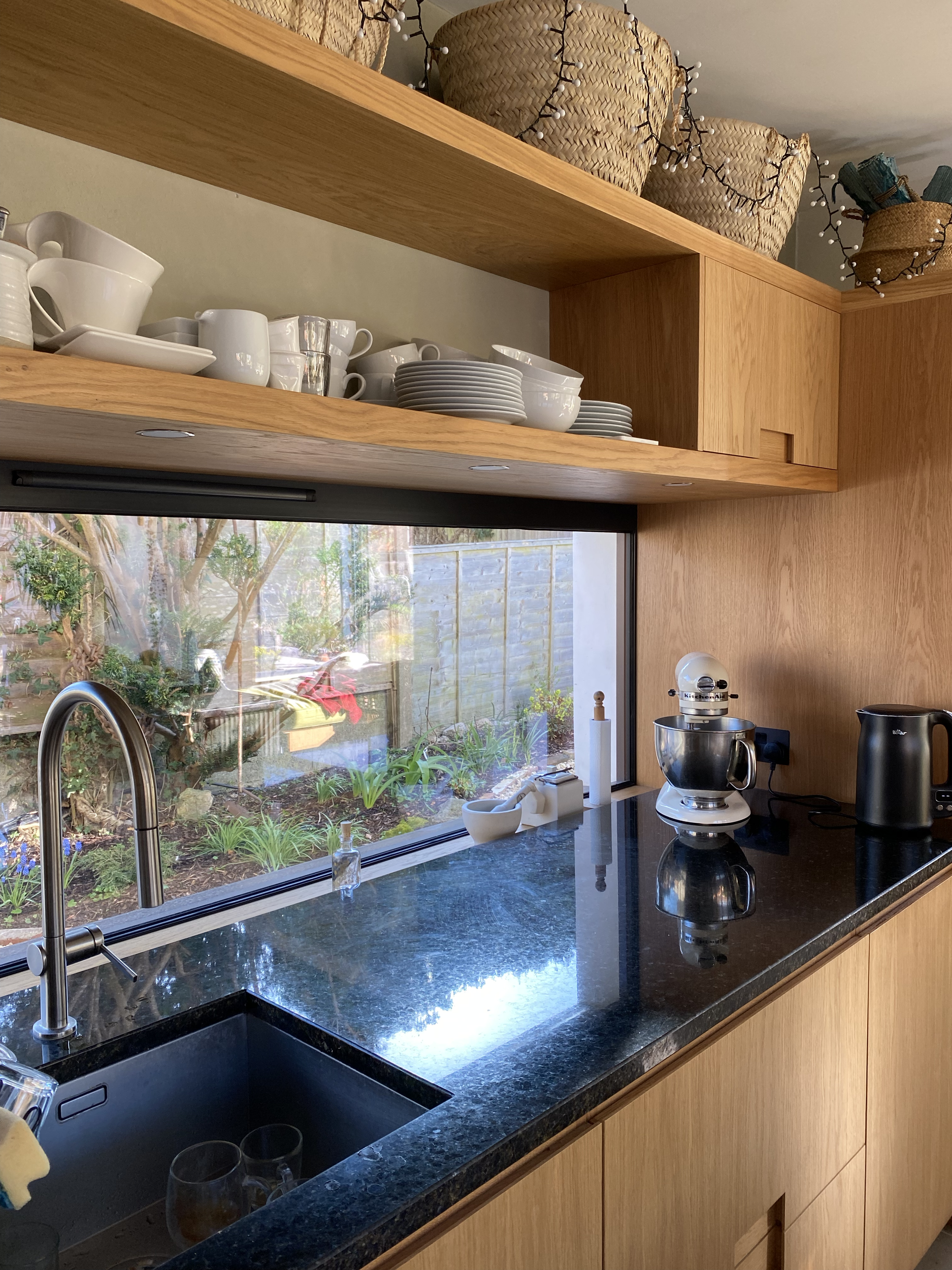 Kitchen view with open shelves containing white dishes and cups, a black countertop with a sink, a window with a garden view, and various kitchen appliances including a stand mixer, toaster, and kettle.