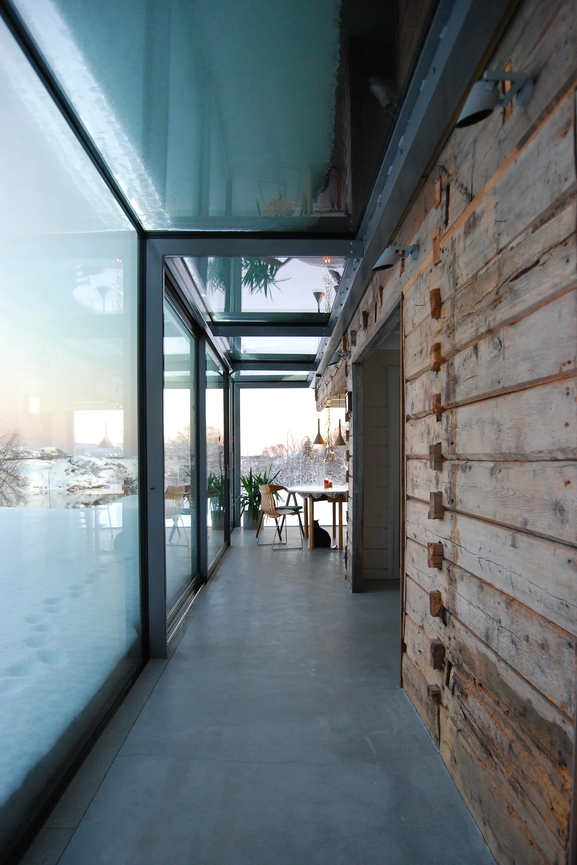 Tilbygg av glass som skaper en glassgang med betonggulv på siden av et gammelt tømmerhus. Kontrast mellom gammelt og nytt.