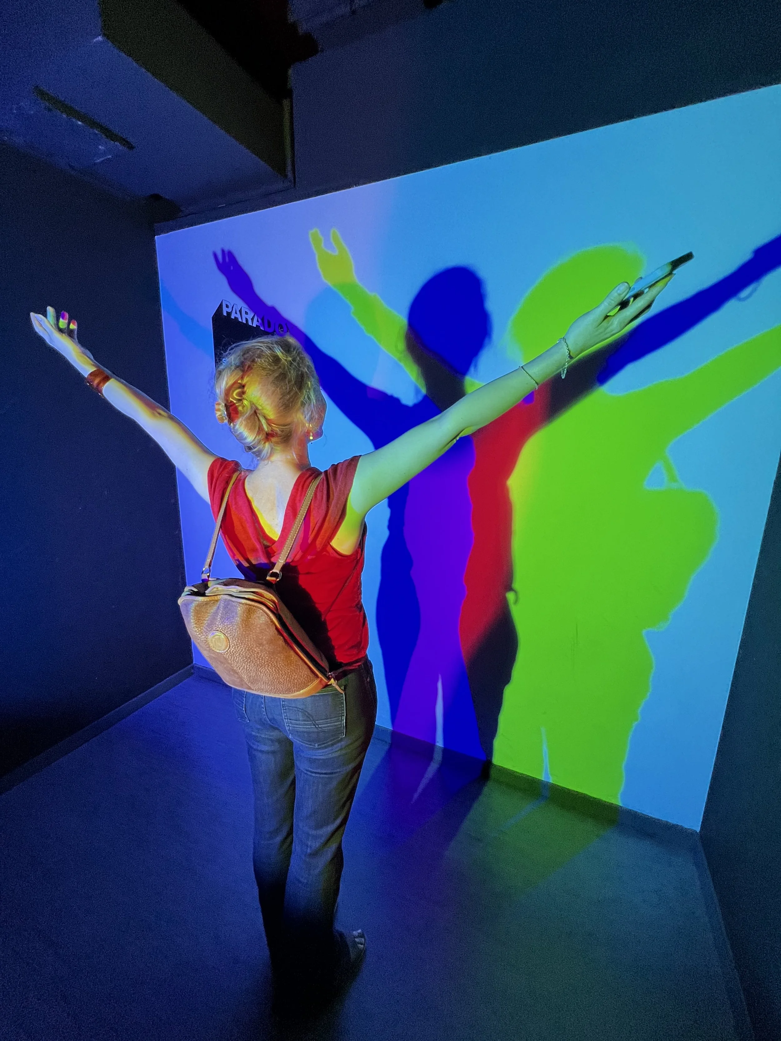 A person in a red shirt and jeans standing with arms outstretched in front of a colorful shadow wall display.