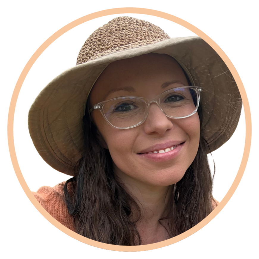 A woman with long dark hair wearing glasses, a wide-brimmed straw hat, and a tan outfit, smiling at the camera.
