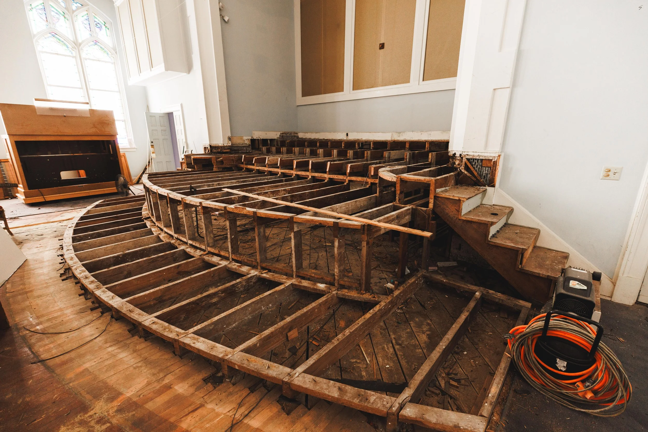 Interior of a building under renovation with wooden framework of a staircase and damaged flooring, construction equipment and orange extension cords on the floor.