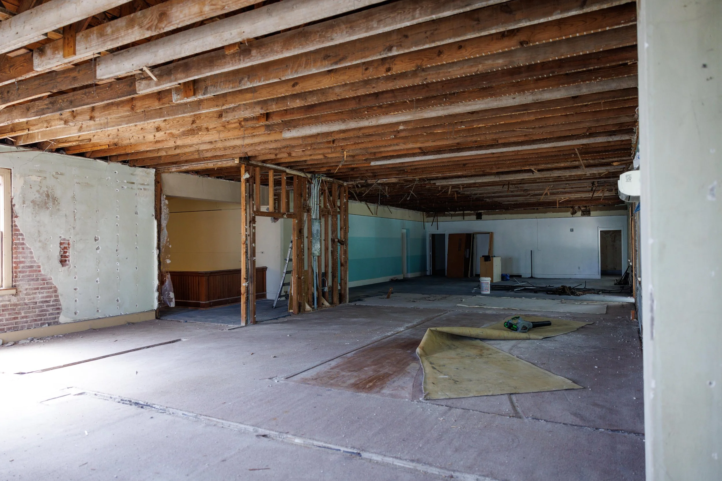Interior of a building under renovation showing exposed wooden ceiling beams, partially demolished walls, construction tools, and materials on the floor.