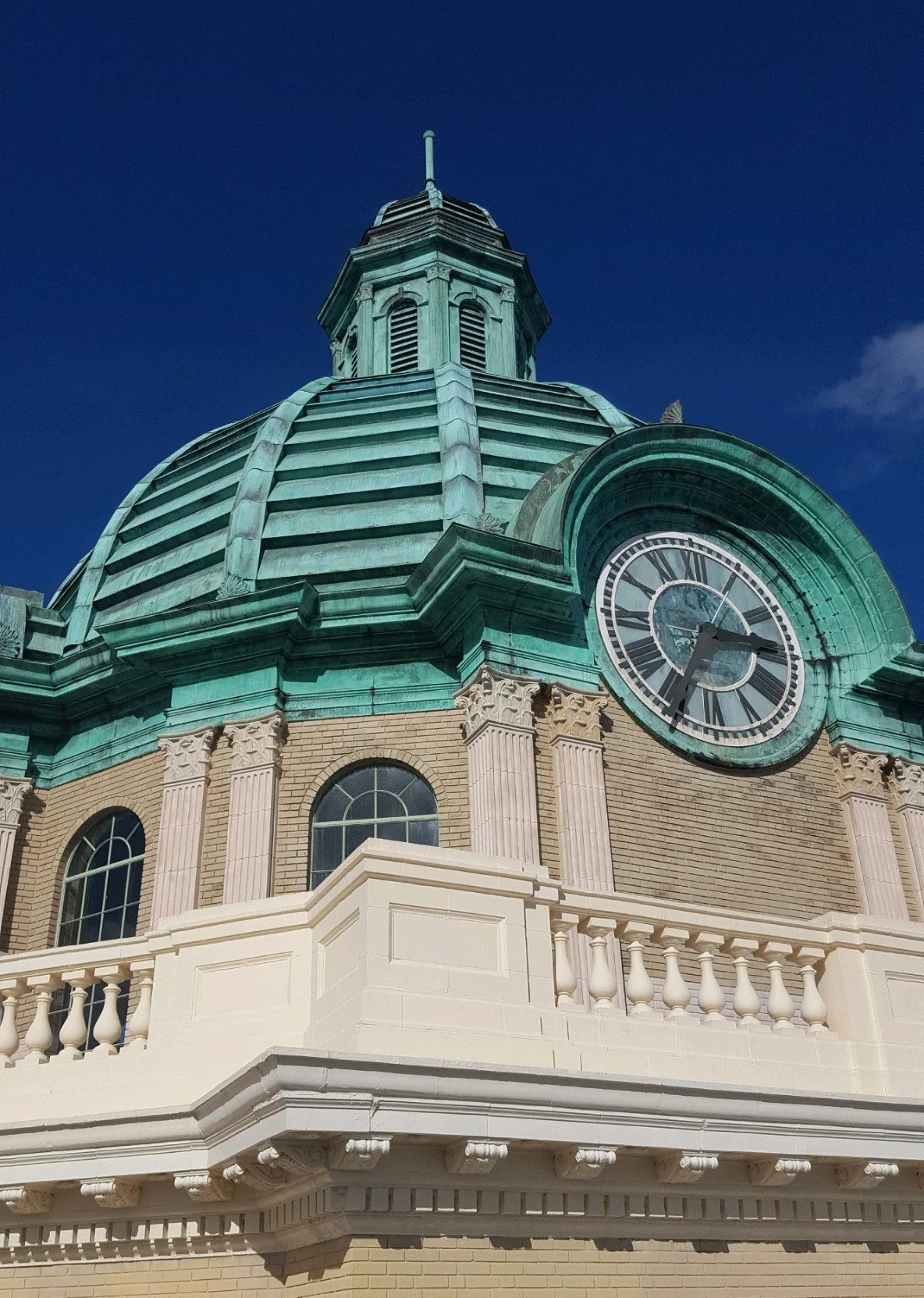 A historic building with a green clock tower and a large clock face showing approximately 2:14, featuring architectural details like a domed roof, columns, and a white balustrade, against a clear blue sky.