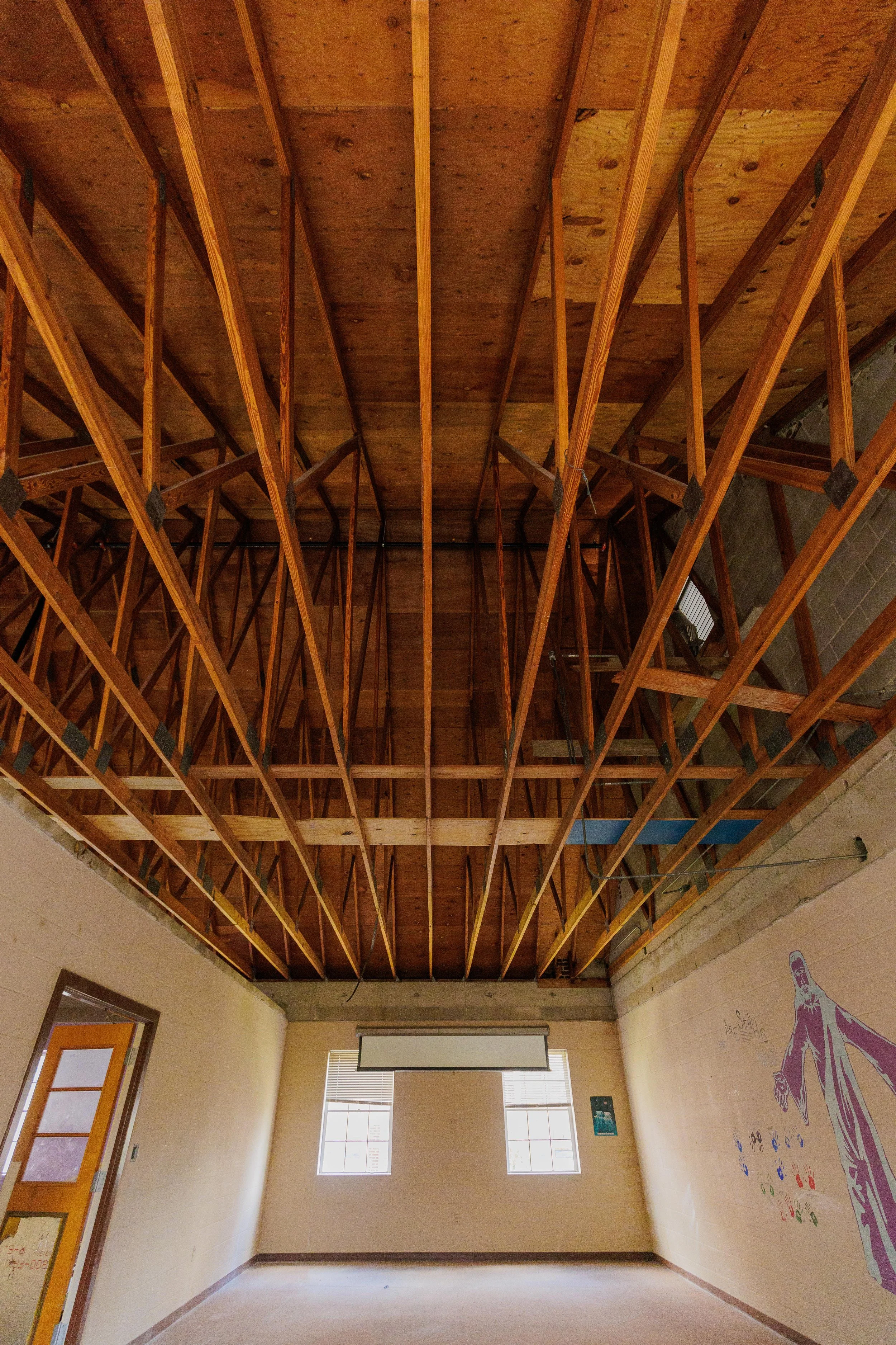 An indoor room with a ceiling under construction, showing exposed wooden framing and unknown structures. The room has two windows on the far wall and a projection screen hanging from the ceiling.