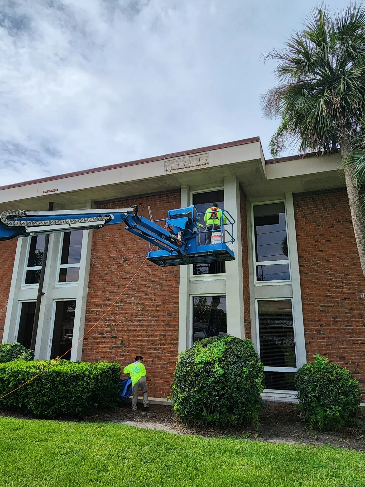 Worker in a lift inspecting or repairing a window of a brick building, with another worker at ground level.