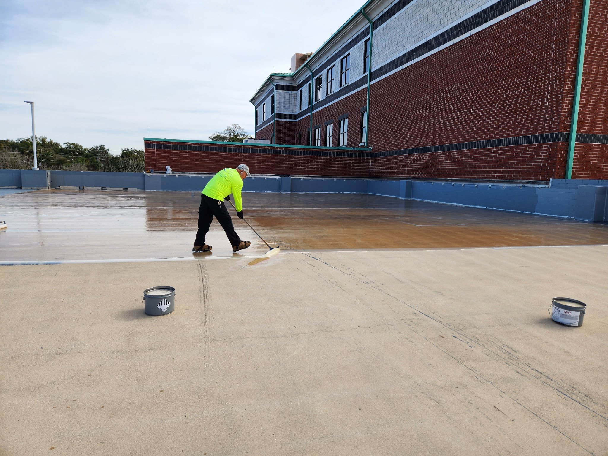 A worker in a bright green safety shirt using a squeegee to apply a coating on a flat rooftop surface, with buckets of coating nearby and a red brick building in the background.