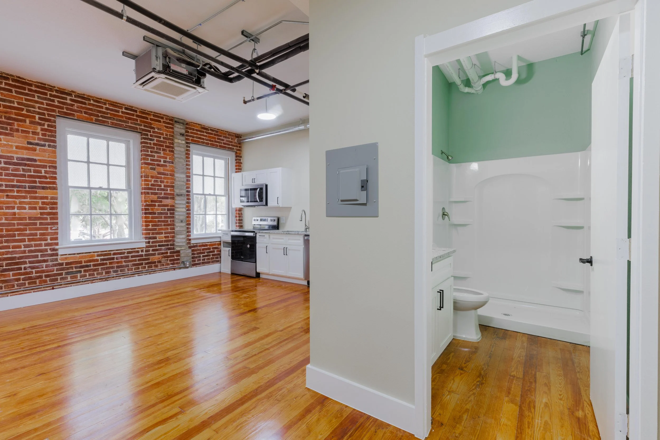 Interior of an apartment with an exposed brick wall, hardwood floors, and large windows. There is a small kitchen area with white cabinets and appliances. A separate bathroom with a toilet and shower stall is visible.