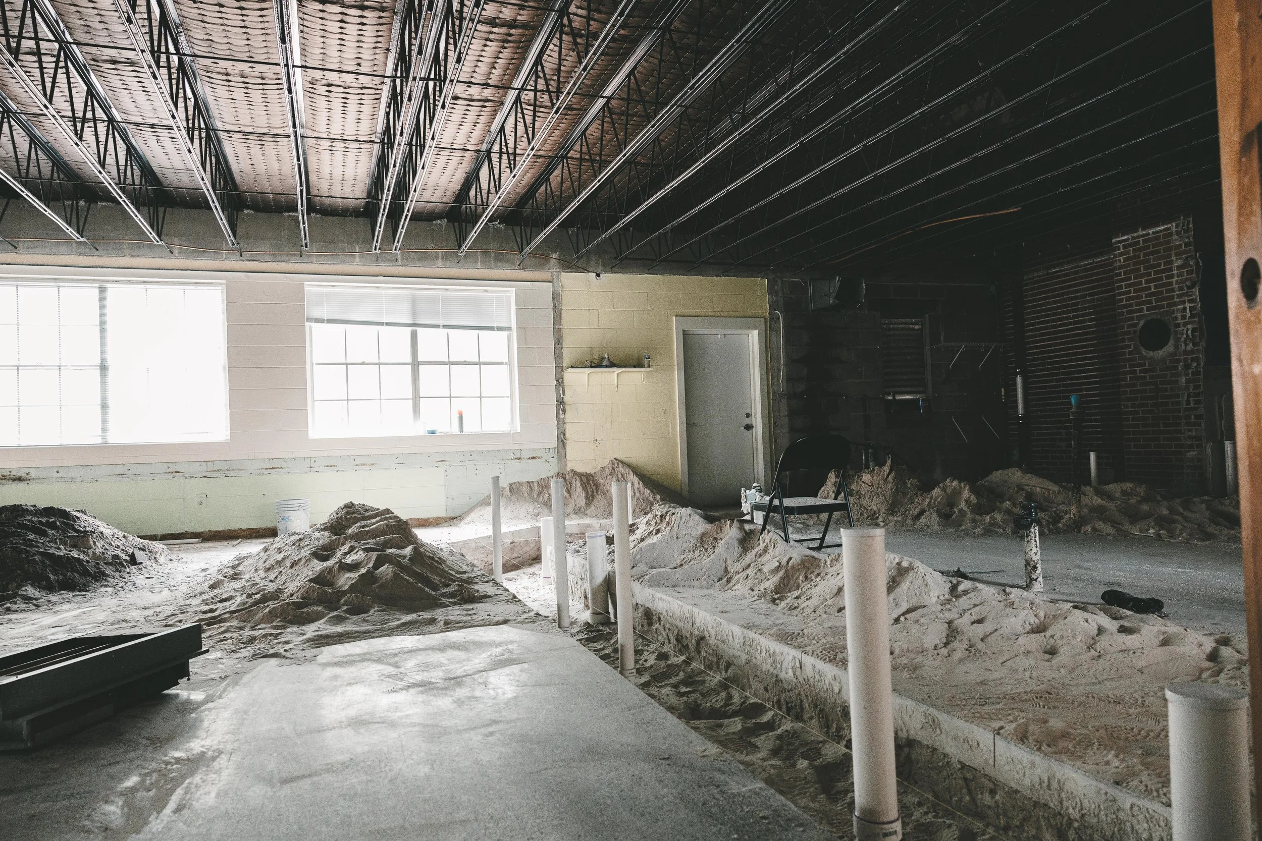 Construction site indoor with piles of sand, pipes, a chair, and construction materials under open ceiling beams, with sunlight coming through windows.