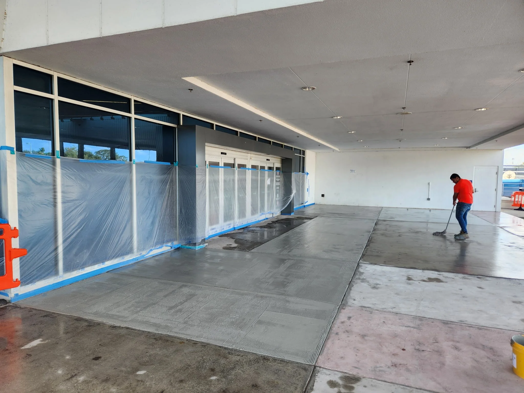A worker in a red shirt and jeans is sweeping the concrete floor outside a building with glass windows, plastic sheeting, and construction barriers.