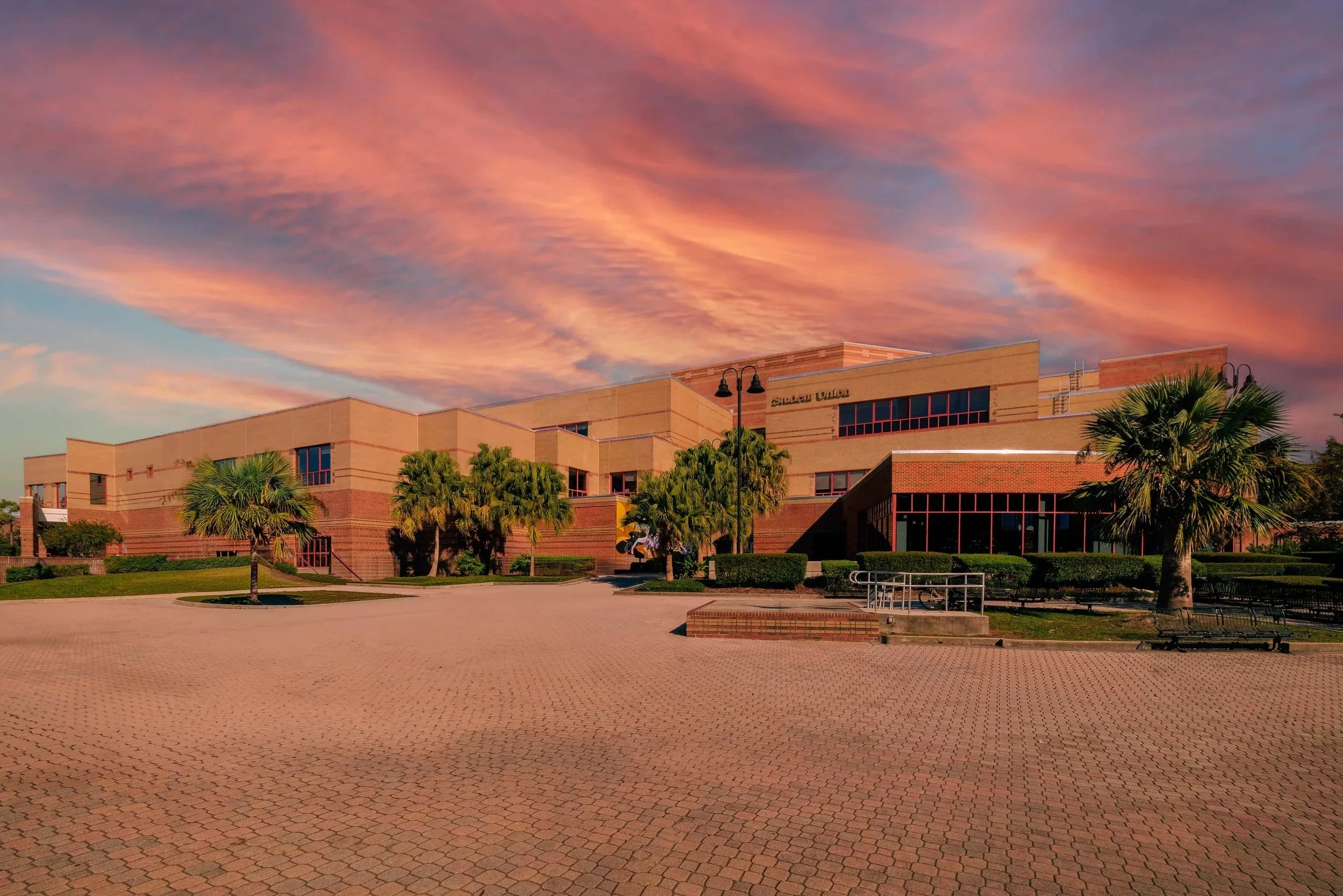 A large brick building with multiple levels and windows, surrounded by palm trees and a paved courtyard, under a colorful sunset sky.